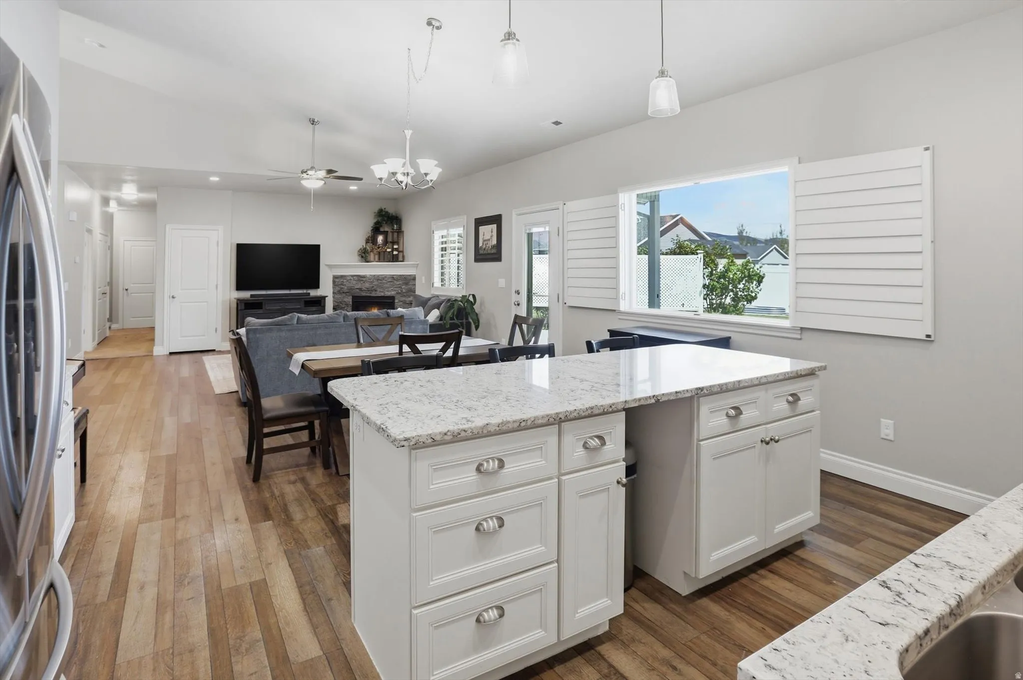 Open-concept living space with wood-finish flooring, a kitchen island featuring a light-toned countertop, and a fireplace with a stone surround