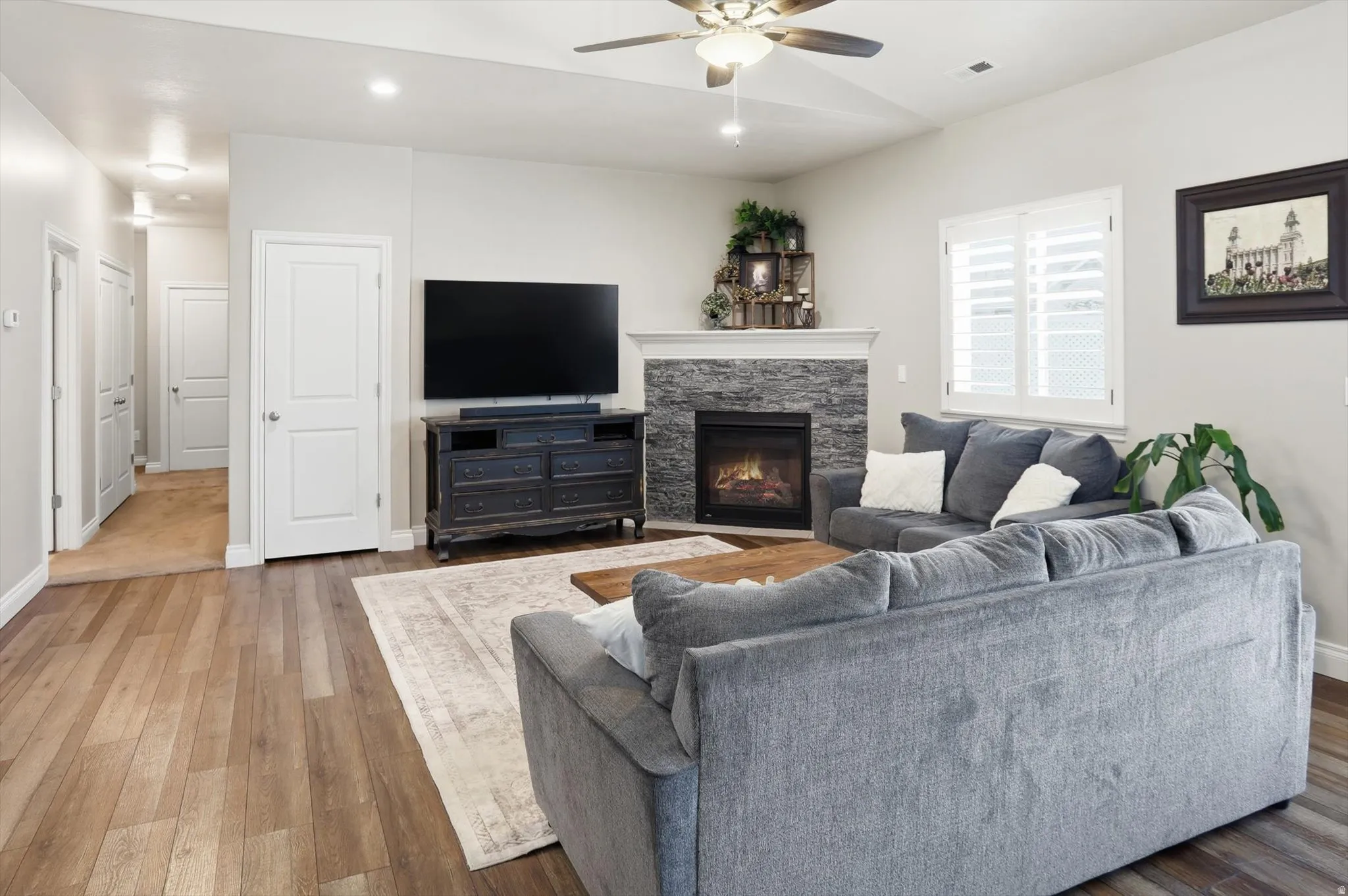 Living space featuring wood-finish flooring, a stacked stone fireplace with a white mantel, and ceiling fan