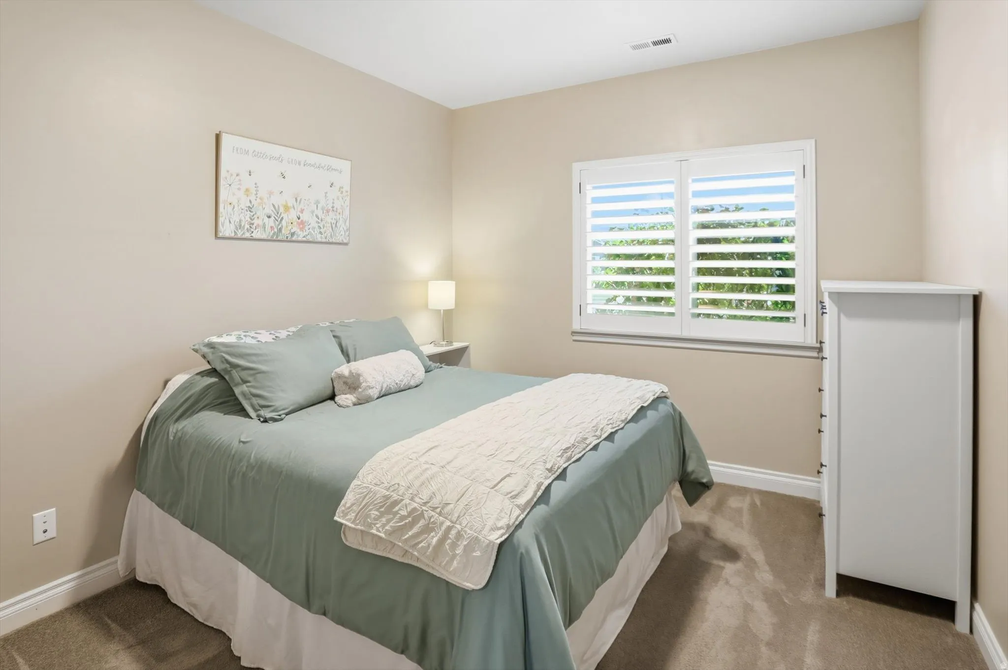 Neutral-toned bedroom featuring a window with white plantation shutters, light beige wall paint, and a textured carpeted floor