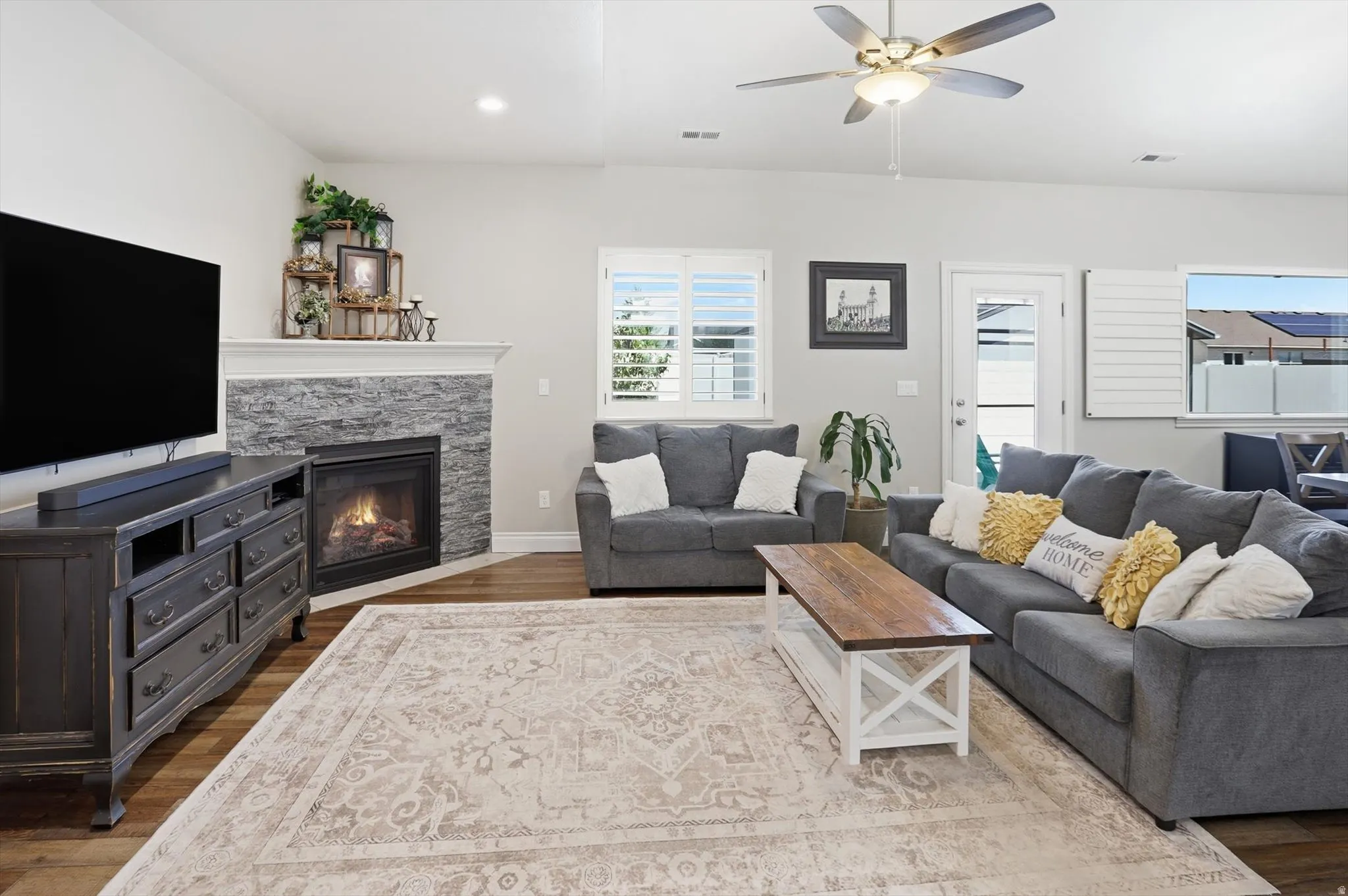 Living area featuring wood-finish flooring, a corner fireplace with stone surround and white mantle, and a ceiling fan with integrated lighting