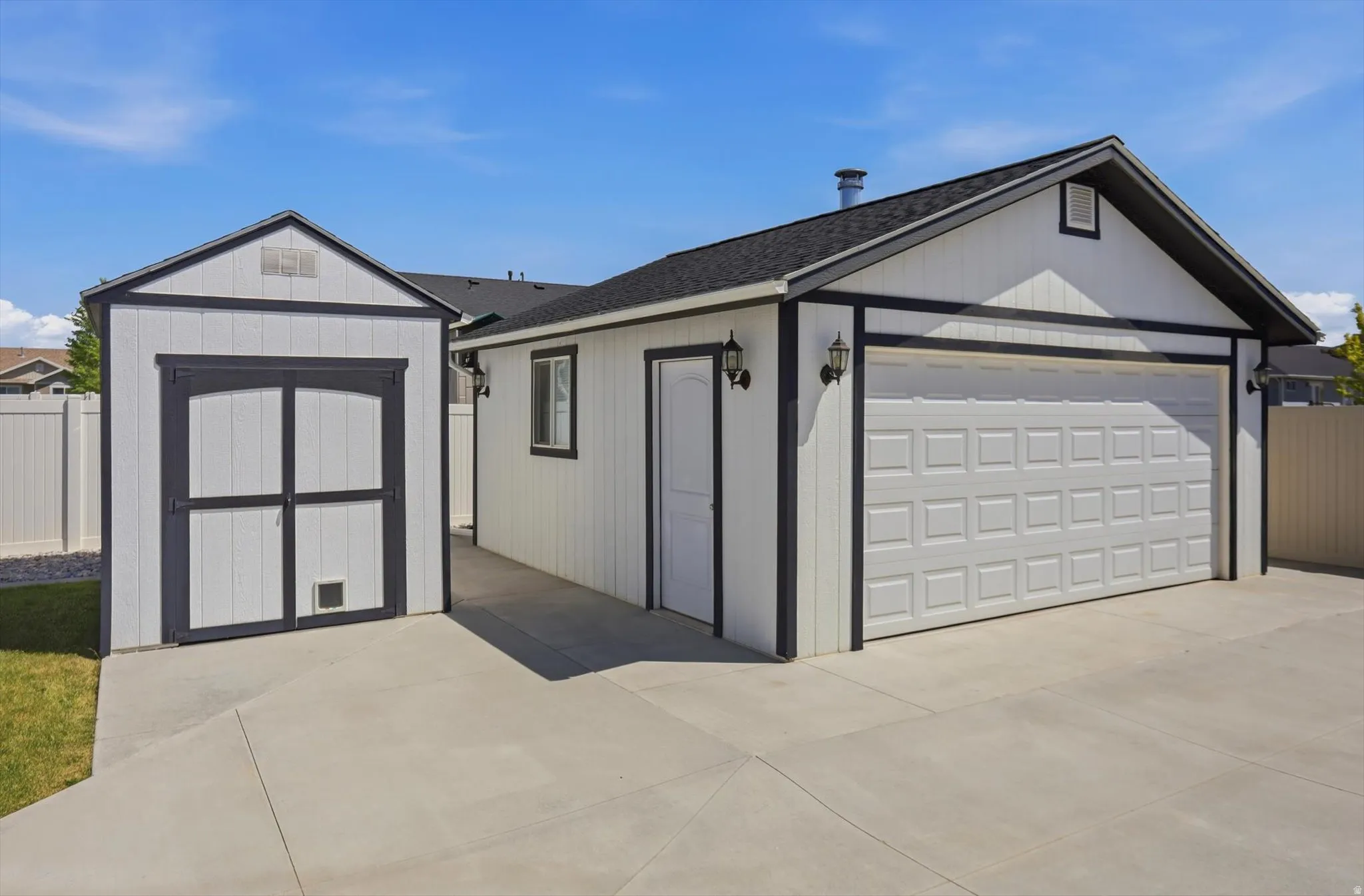 Detached garage with white paneling and dark trim, featuring a paneled garage door and exterior lighting fixtures