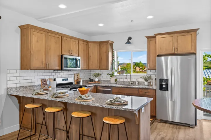Kitchen featuring wood-finish cabinetry, stainless steel appliances, speckled stone countertops, a subway tile backsplash, and an island with bar seating