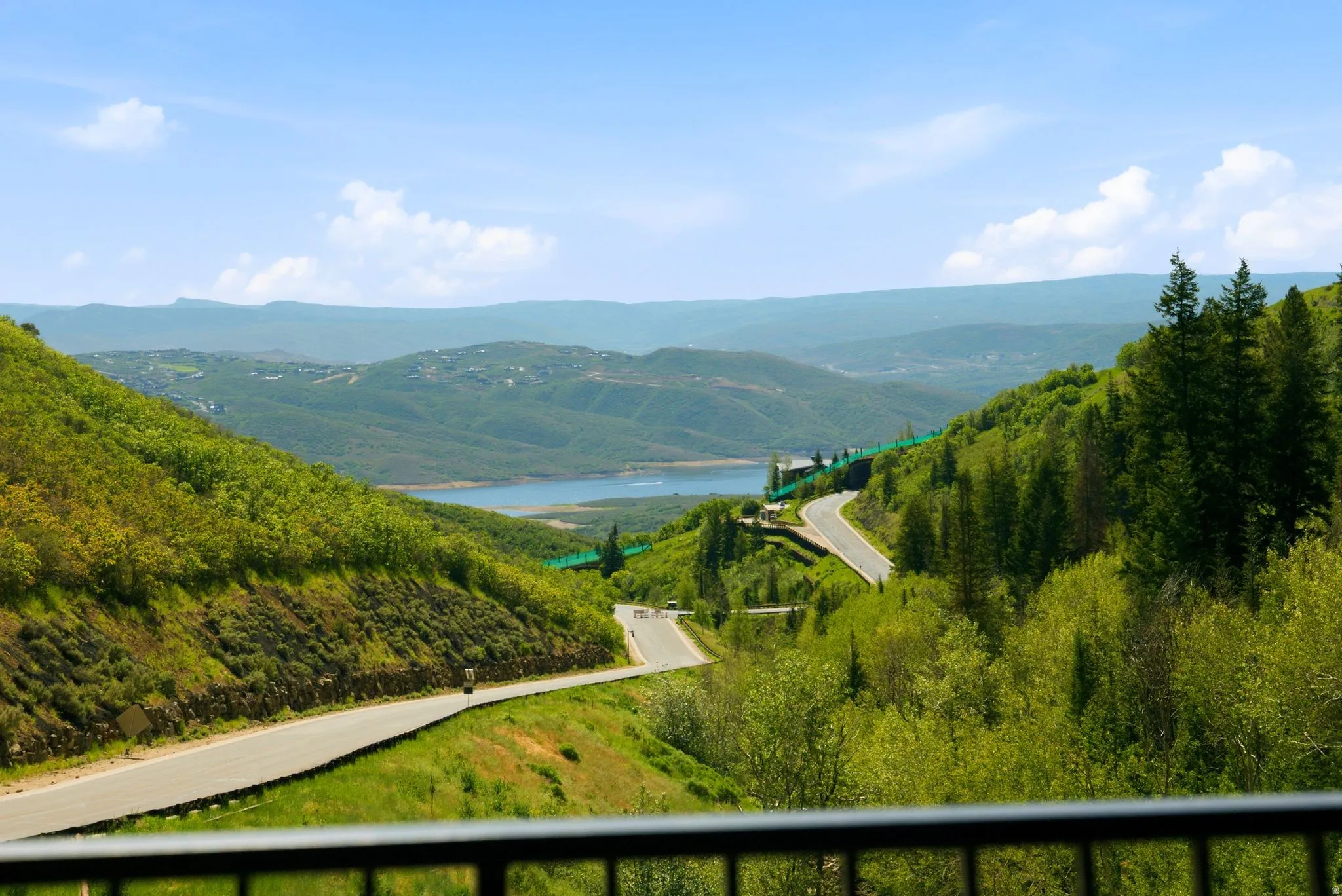Expansive mountain and lake scenery, featuring winding roads, a clear blue sky, distant hills, and abundant green foliage