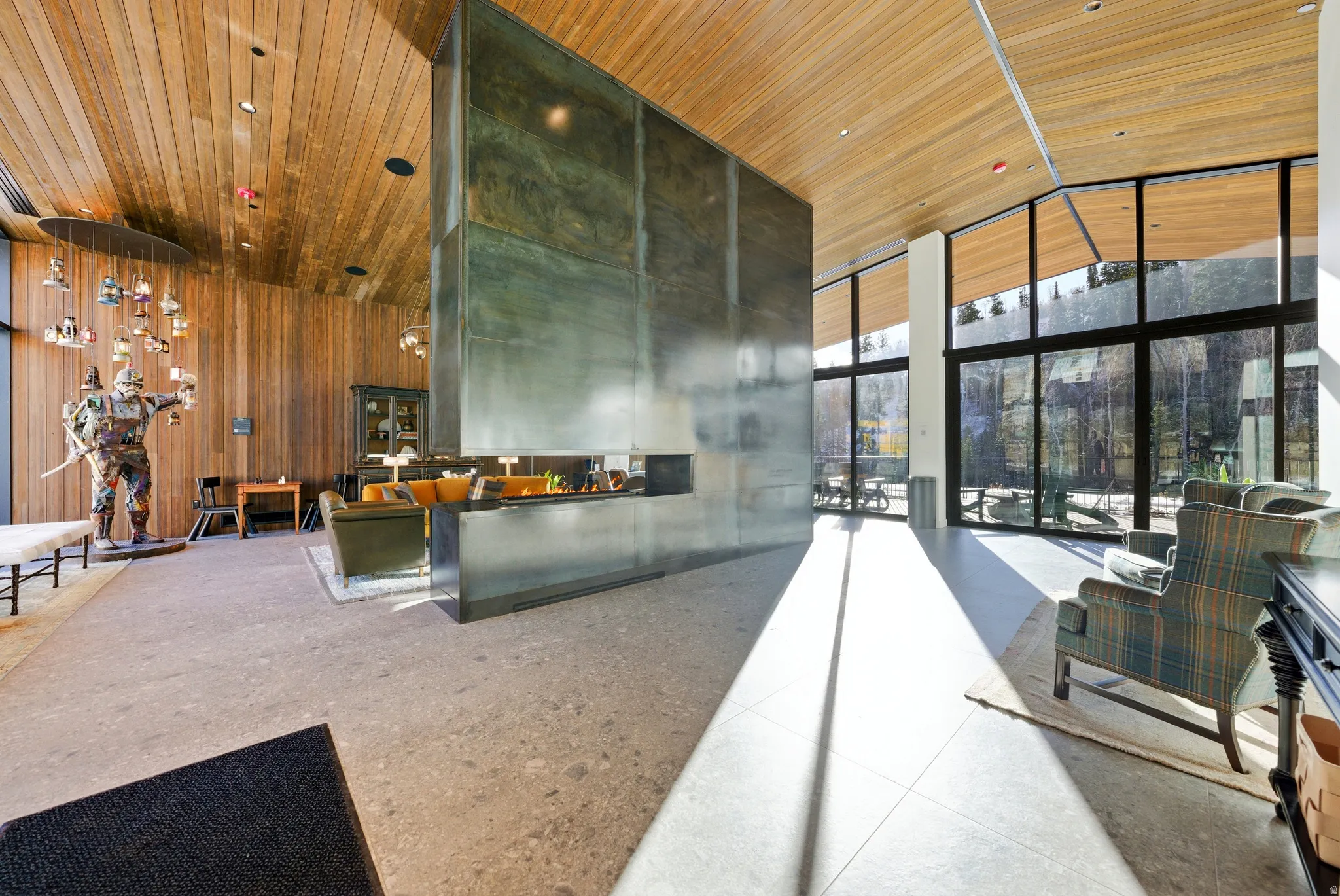 Spacious interior featuring a large metal fireplace, wood-paneled walls and ceiling, full-height windows, and stone flooring