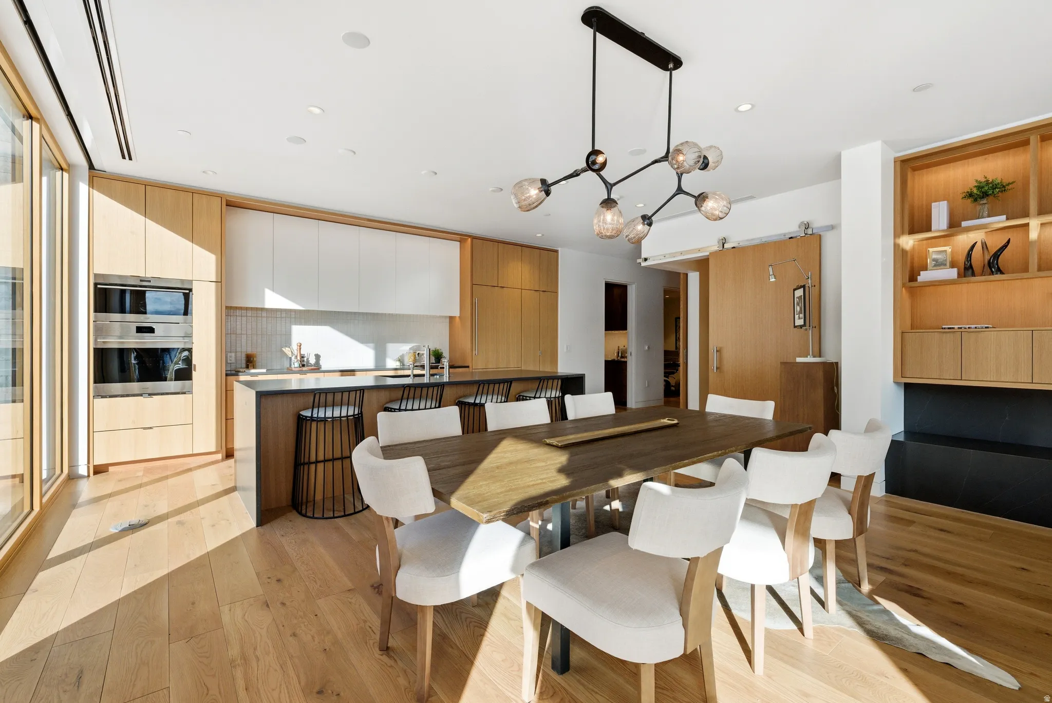 Modern open-concept kitchen and dining area featuring wood-finish flooring, a contemporary linear chandelier, integrated double ovens, and a built-in shelving unit with lower cabinetry