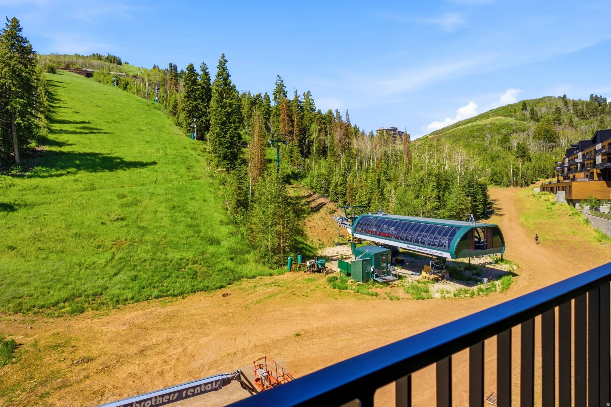 Mountain landscape featuring a ski lift structure, a vibrant green slope, and mature evergreen trees