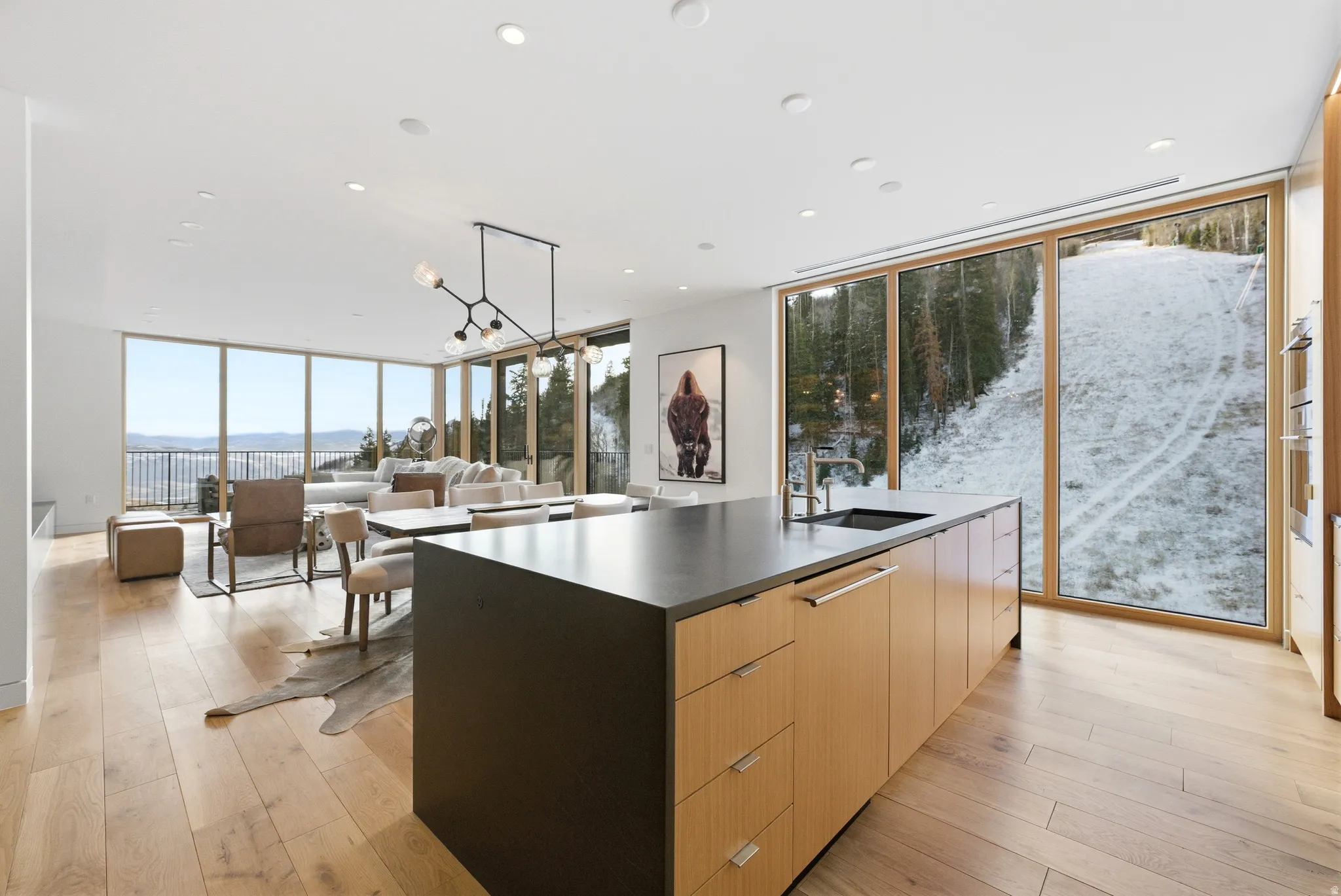 Open-concept living space featuring wood-finish flooring, a contemporary kitchen island with a dark countertop and integrated sink, and expansive floor-to-ceiling windows