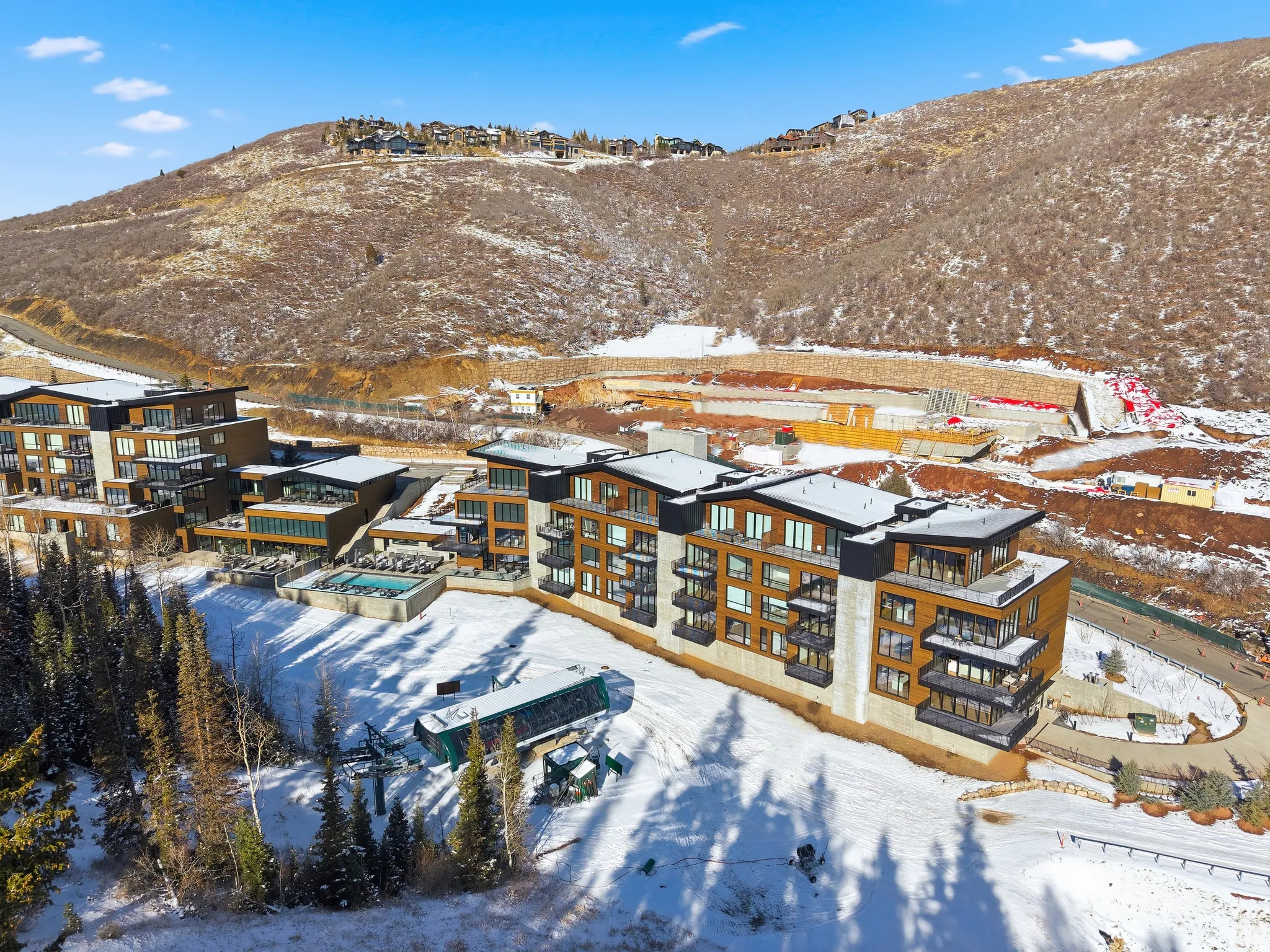 Modern mountain architecture featuring multiple multi-story buildings with balconies, a ski lift, and an outdoor pool area with lounge seating