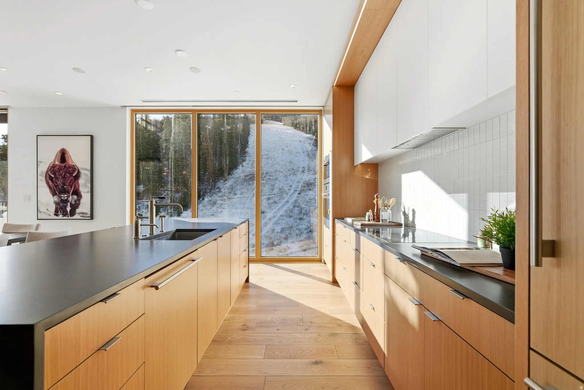 Modern kitchen featuring abundant natural light, full wall-height glass doors showcasing a snow-covered landscape, integrated wood-finish cabinetry, a large kitchen island with an undermount sink, and recessed ceiling lighting