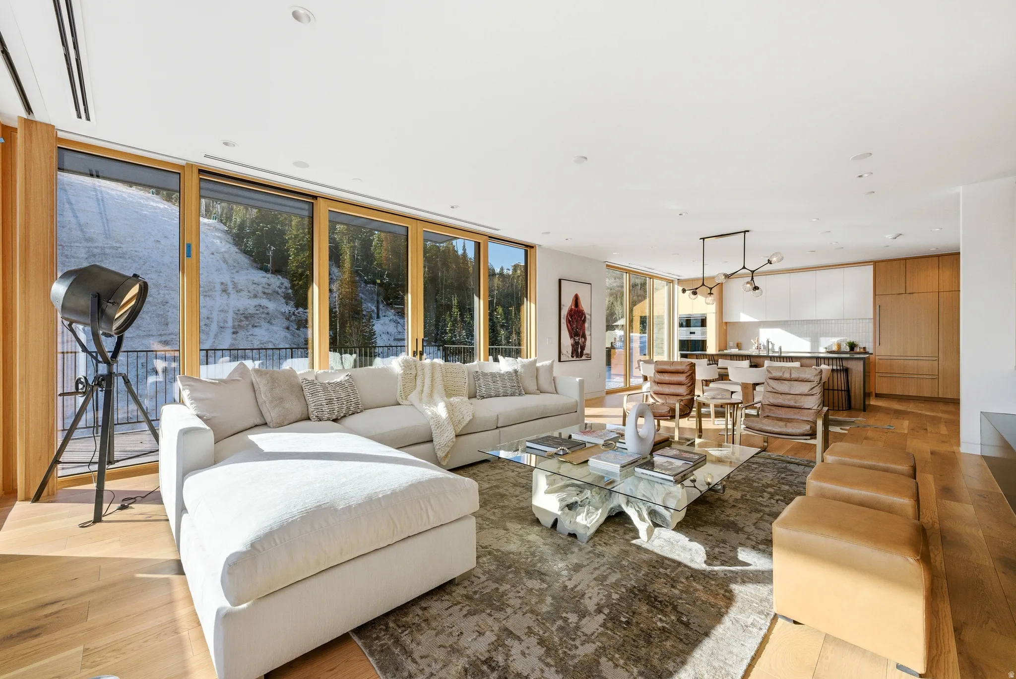 Open-concept living space with expansive floor-to-ceiling glass doors, natural wood-finish flooring, a modern linear chandelier, and integrated kitchen cabinetry