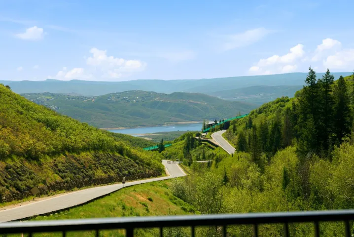 Expansive mountain and lake scenery, featuring winding roads, a clear blue sky, distant hills, and abundant green foliage