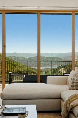 Floor-to-ceiling windows with natural wood frames offer expansive mountain and distant water scenery