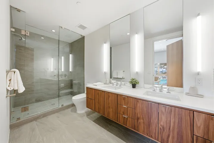 Spacious bathroom featuring a dual vanity with wood-finish cabinetry and white countertops, a frameless glass shower enclosure, and large-format tile flooring