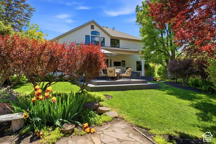 Rear exterior featuring a multi-level deck, covered patio, manicured lawn, mature trees, and landscaping with stone pathways