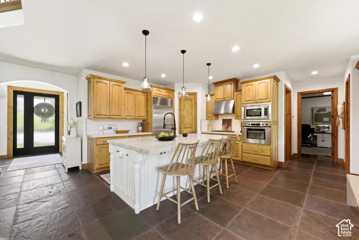 Spacious kitchen featuring light wood cabinetry, stainless steel appliances, a large center island with seating, and dark slate tile flooring