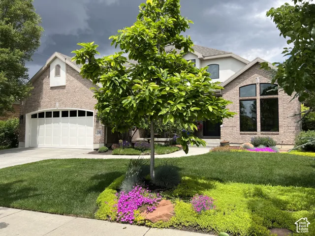 Brick facade with arched windows, integrated two-car garage, manicured lawn, and vibrant garden landscaping