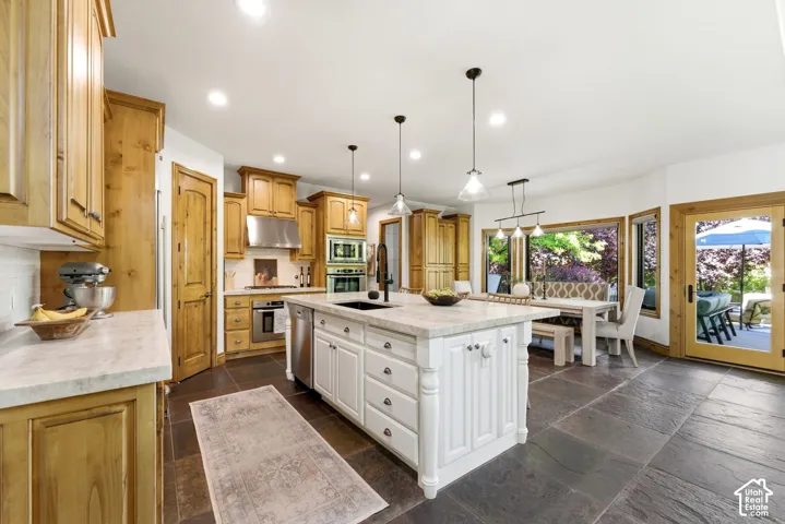 Spacious kitchen featuring a large central island with seating, natural wood cabinetry, stainless steel appliances including a built-in oven and microwave, dark stone-finish flooring, and ample natural light from large windows and a glass-paneled door