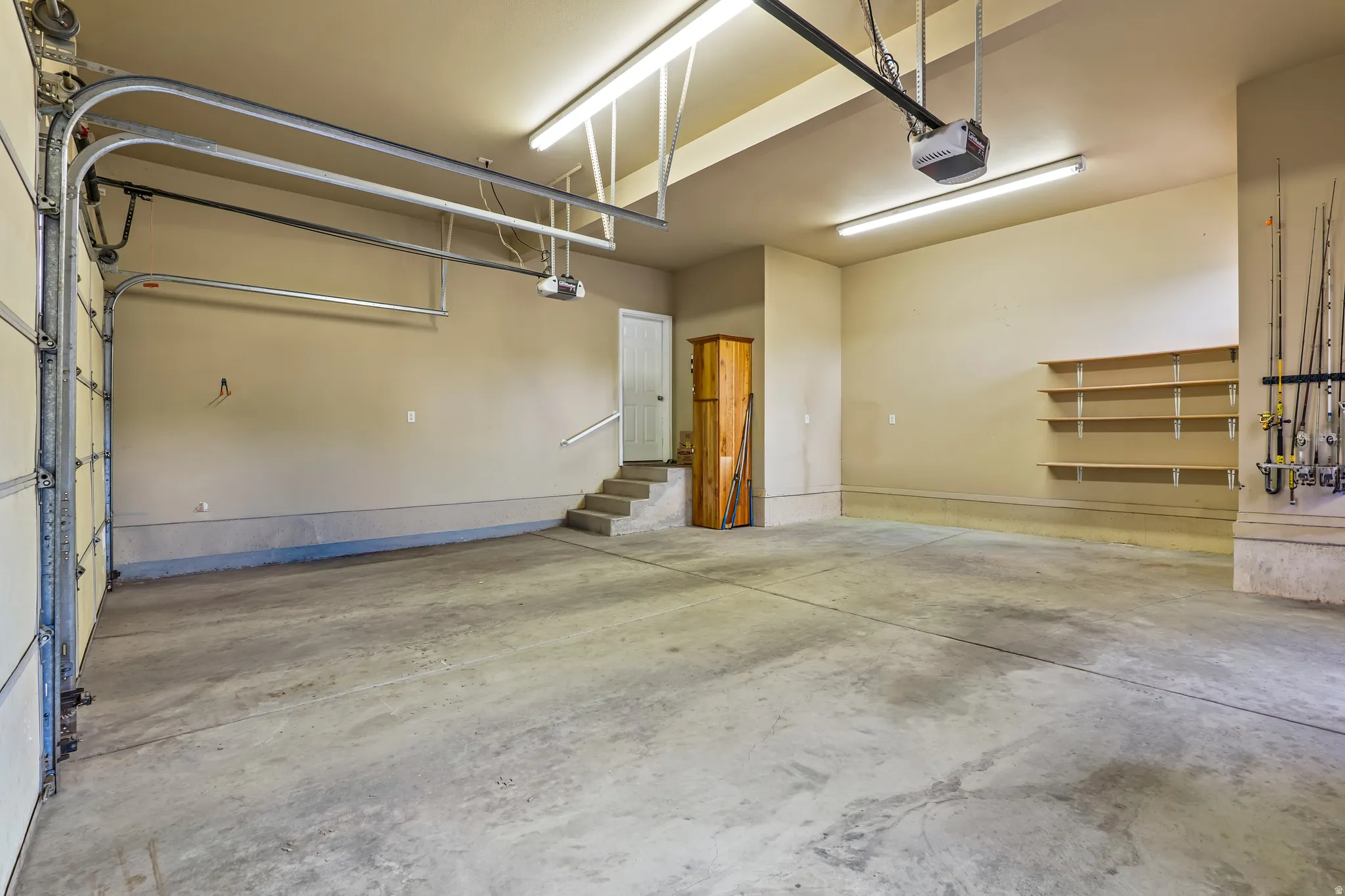 Spacious two-car garage featuring a concrete floor, built-in shelving, overhead fluorescent lighting, and a white interior access door with a handrail