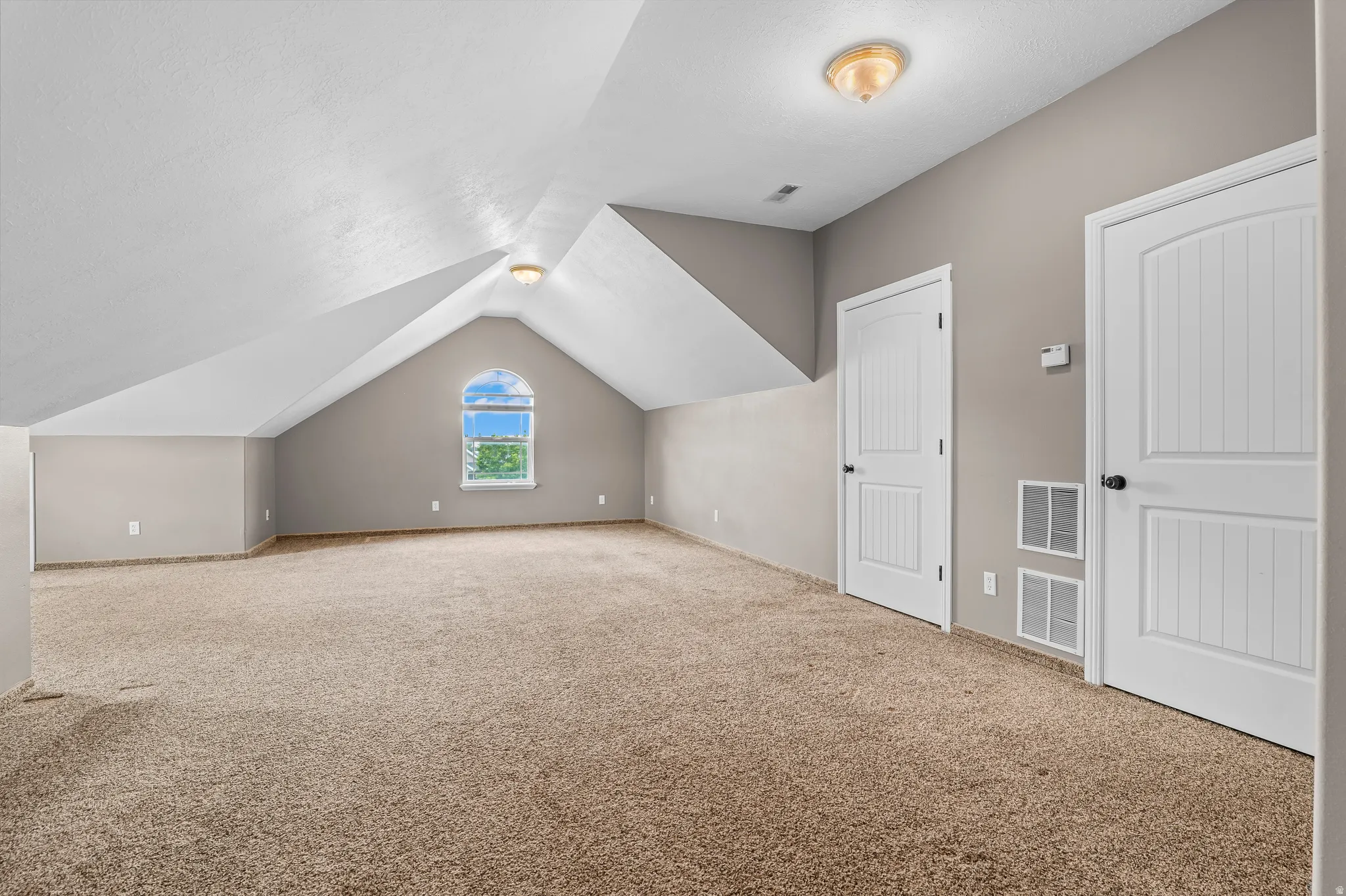Spacious carpeted room featuring a vaulted ceiling with an arched window, two paneled doors, and ceiling-mounted lighting fixtures