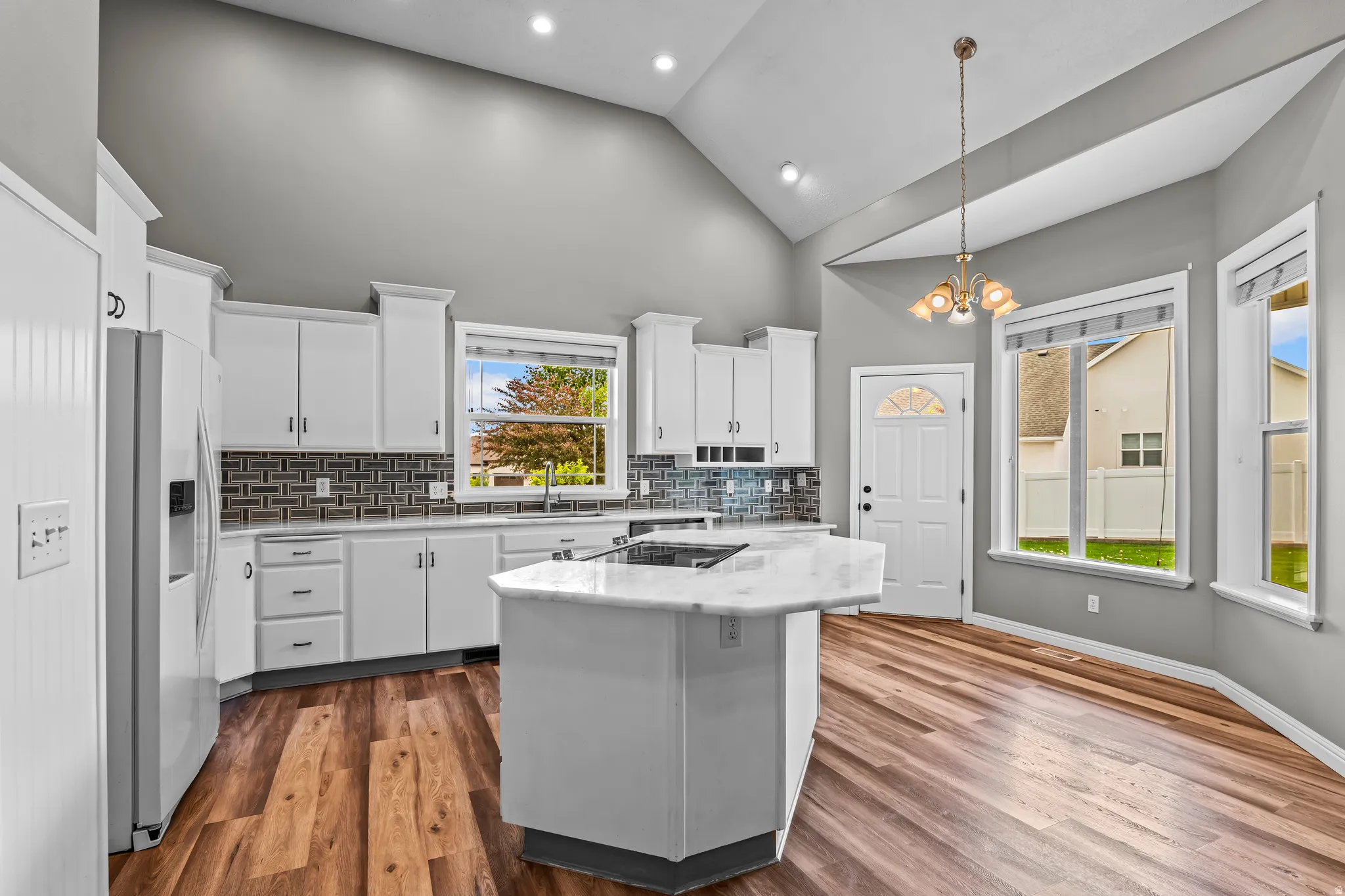 Modern kitchen featuring vaulted ceilings, recessed lighting, wood-finish flooring, a central island with a light-toned countertop, and white cabinetry with dark hardware