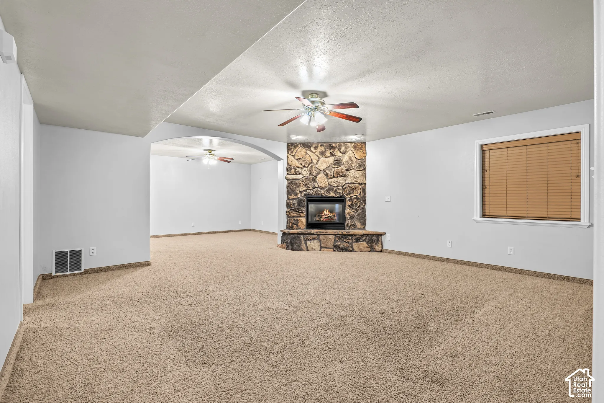 Expansive carpeted room featuring a stone fireplace with hearth, a ceiling fan, and a single window with blinds