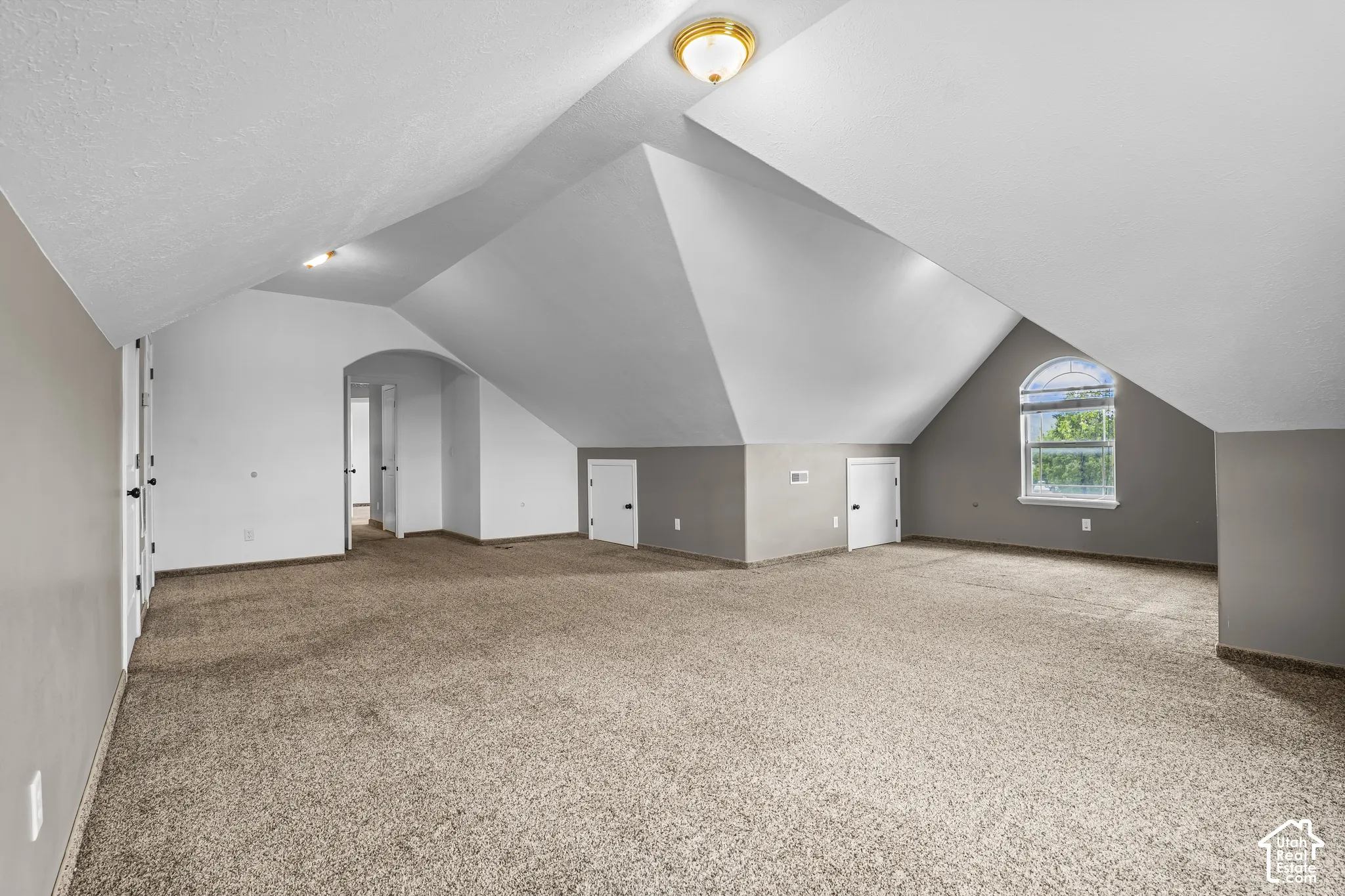 Spacious room featuring vaulted ceilings, neutral-toned carpeting, a prominent arched window, and multiple interior doors