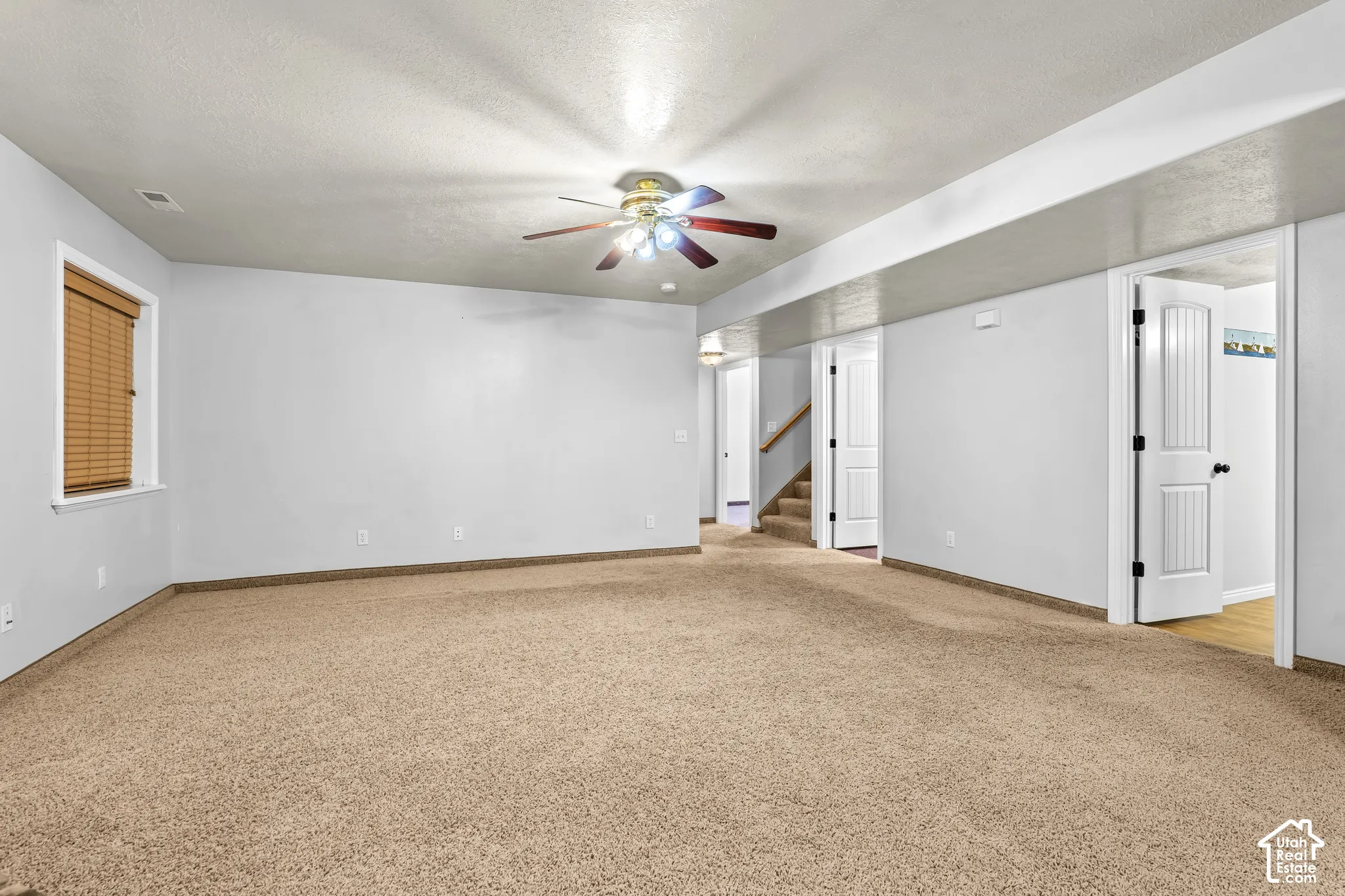 Spacious carpeted room featuring a ceiling fan with integrated lighting, a window with wood-finish blinds, and multiple doorways providing access to other areas of the property