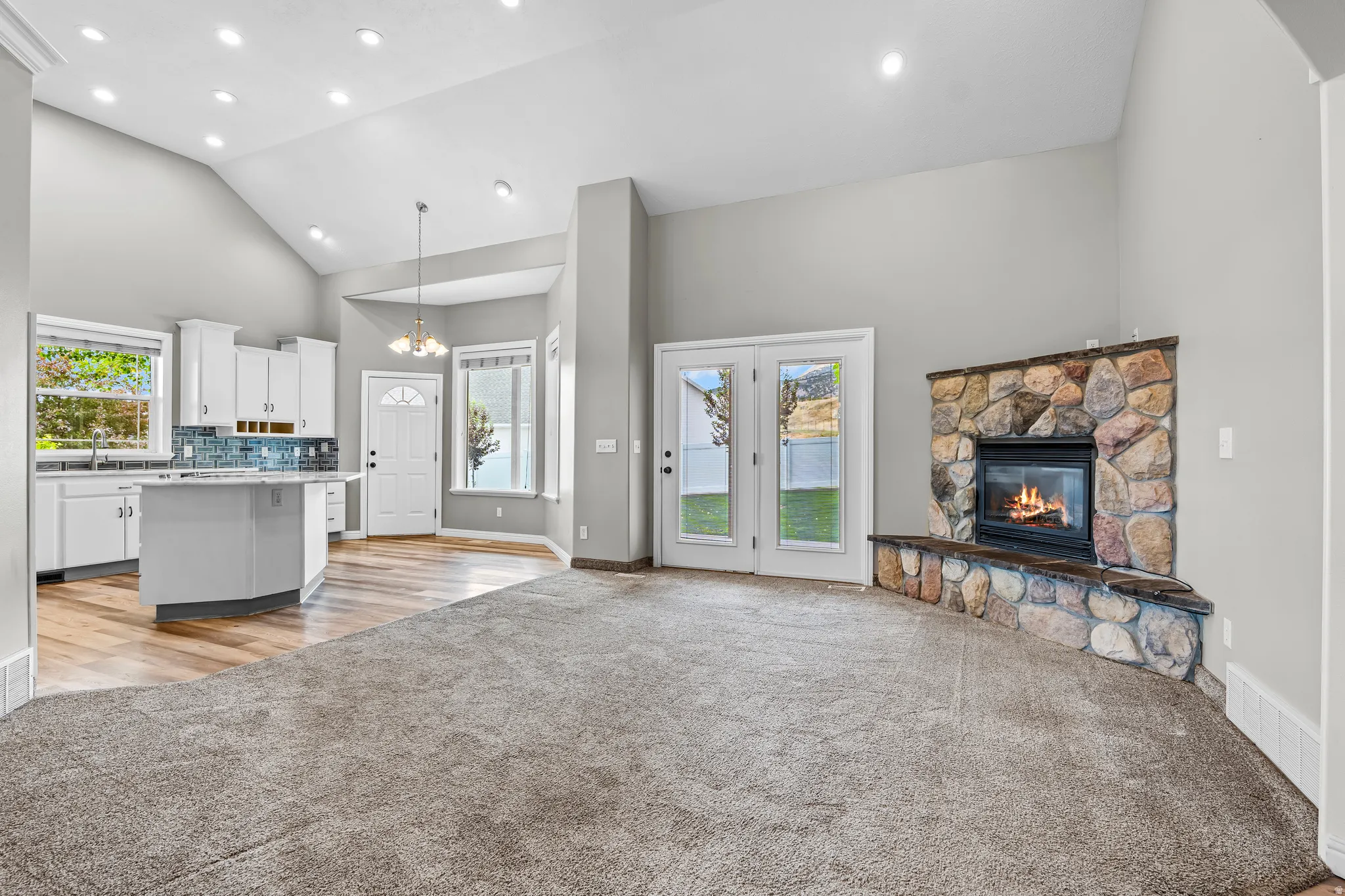 Spacious living area featuring a stone-clad fireplace and hearth, French doors, vaulted ceilings with recessed lighting, a kitchen island with seating, and wood-finish flooring