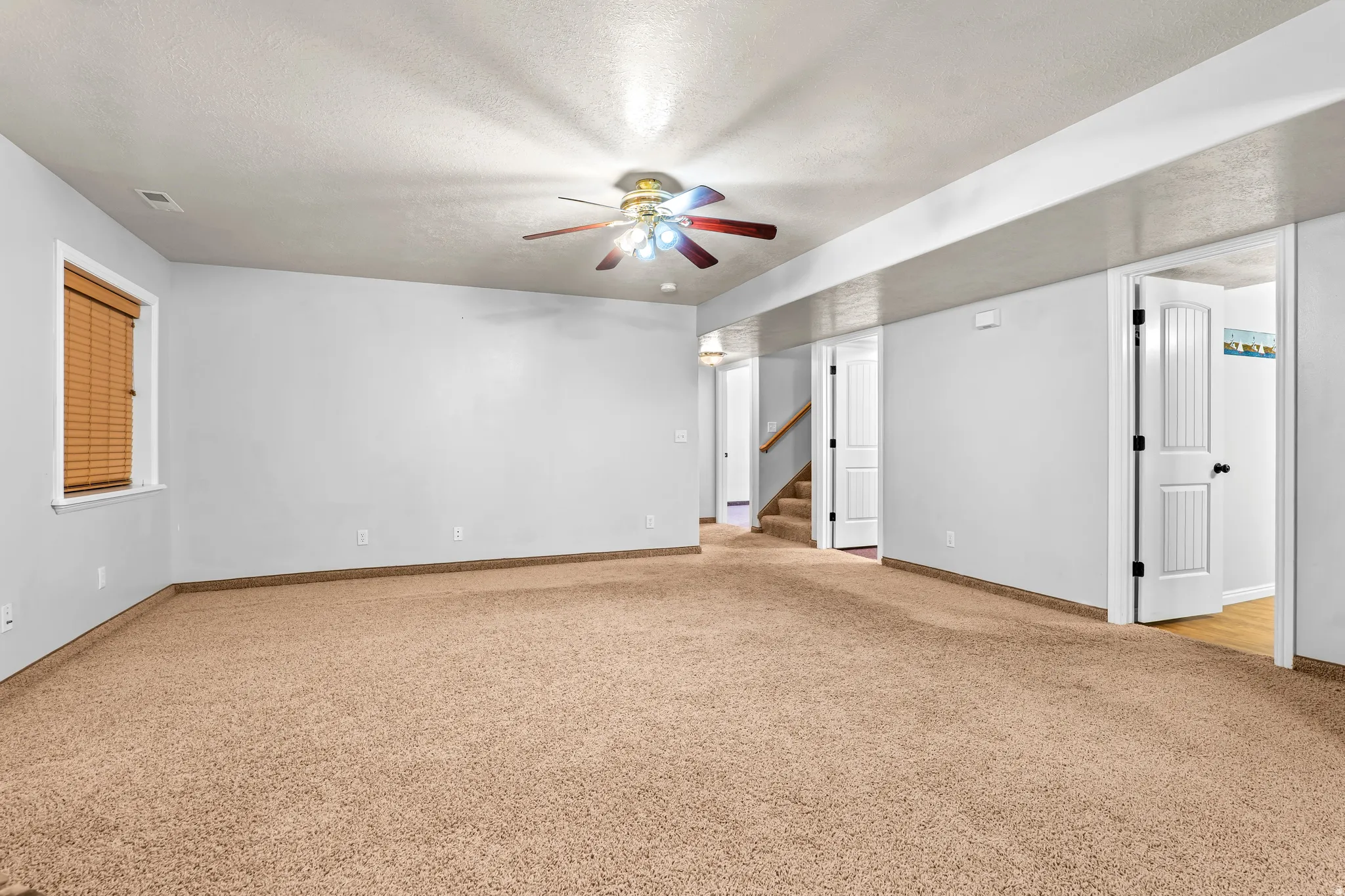 Spacious carpeted room featuring a ceiling fan with integrated lighting, a window with wood-finish blinds, and multiple doorways providing access to other areas of the property