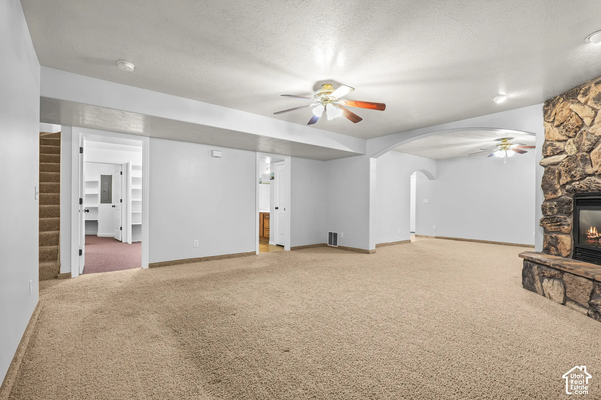 Spacious carpeted room featuring a stone-clad fireplace with a hearth, multiple ceiling fans, and an arched doorway leading to an additional space