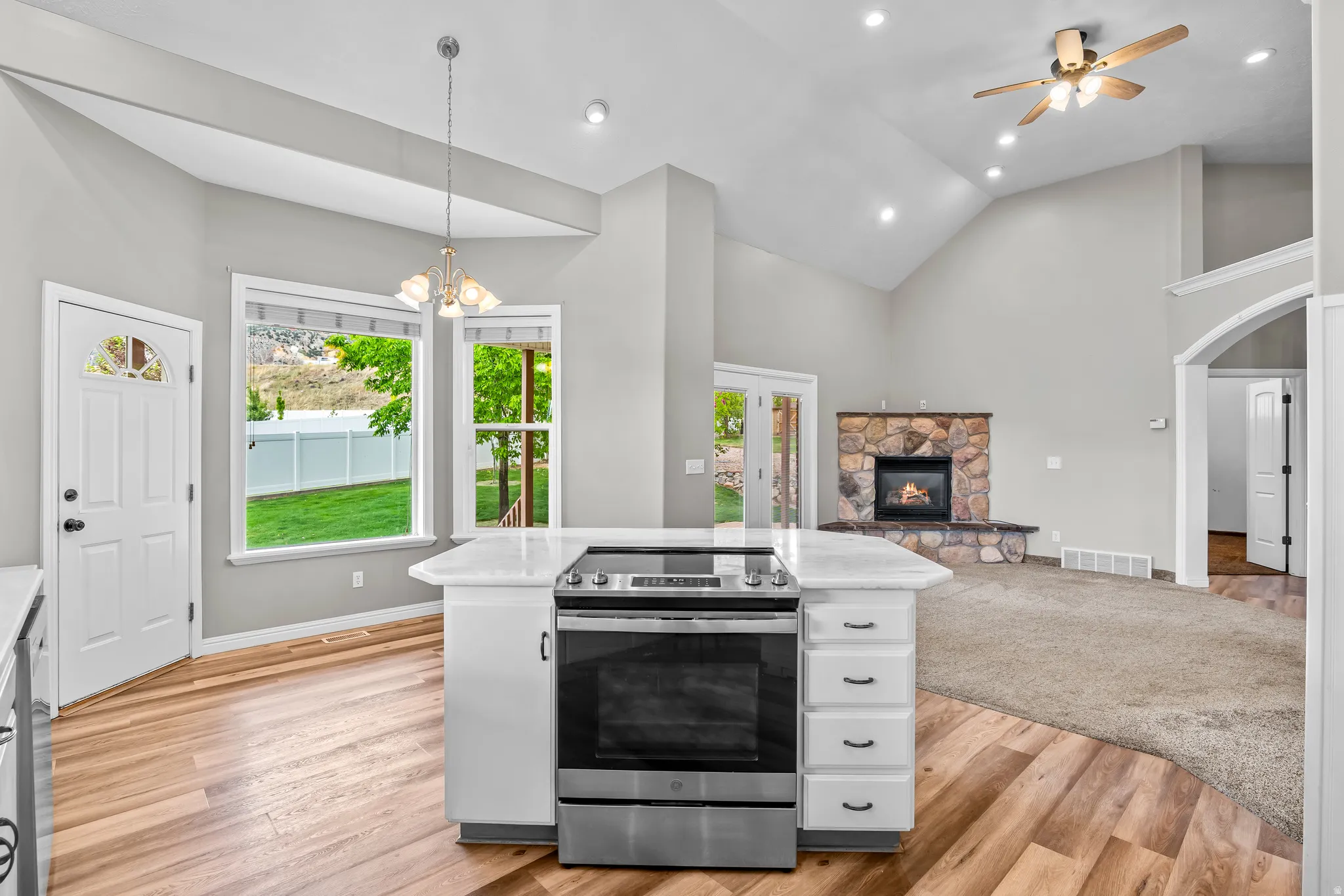 Open-concept living area featuring a kitchen island with built-in stainless steel range, light wood-finish flooring, and a stone-clad fireplace