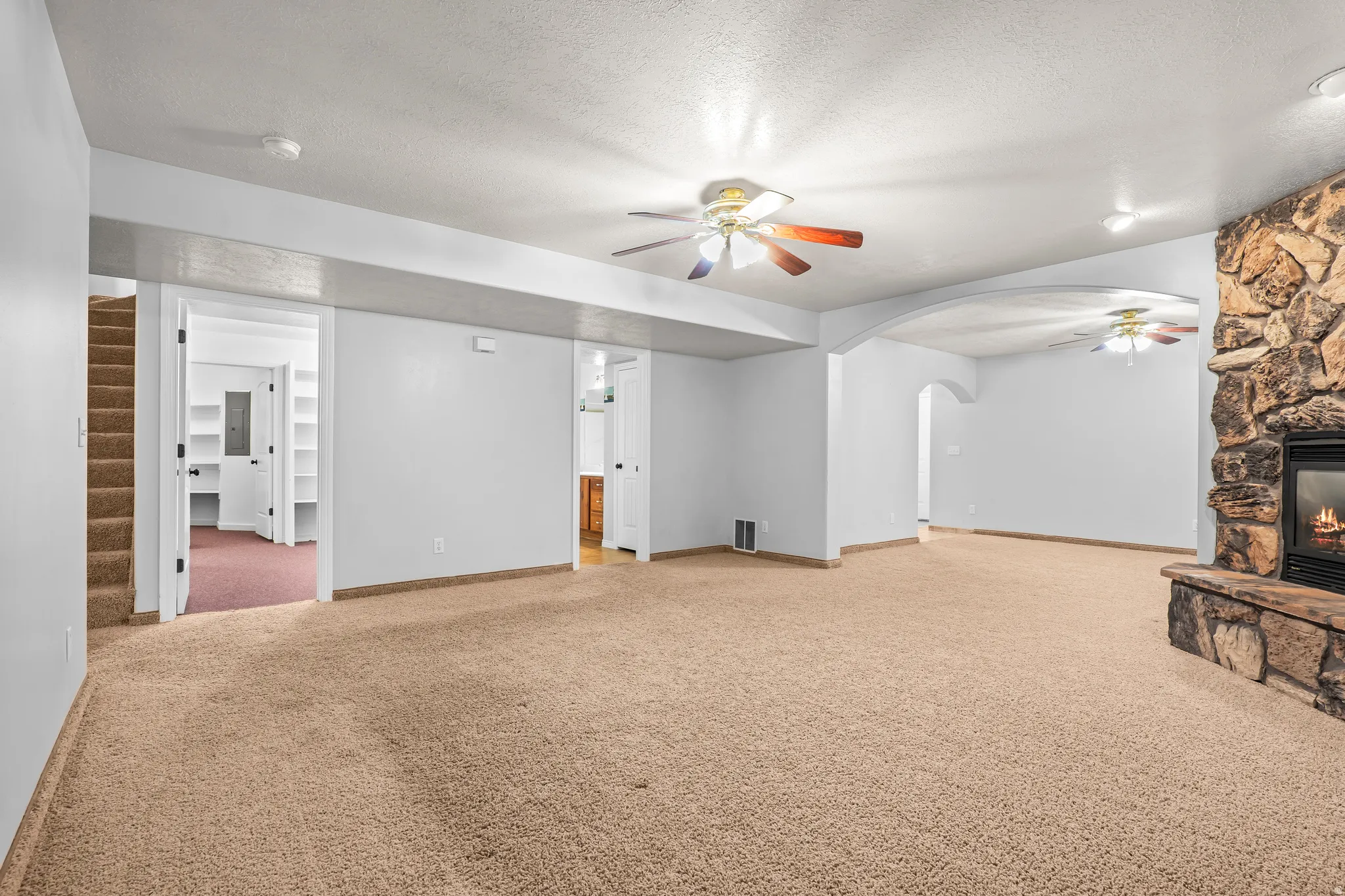 Spacious carpeted room featuring a stone-clad fireplace with a hearth, multiple ceiling fans, and an arched doorway leading to an additional space