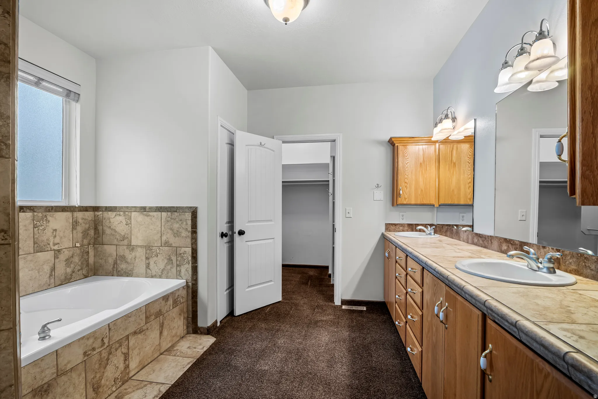 Bathroom featuring a built-in soaking tub with tile surround, a dual vanity with light-colored countertops and wood cabinetry, and an overhead lighting fixture