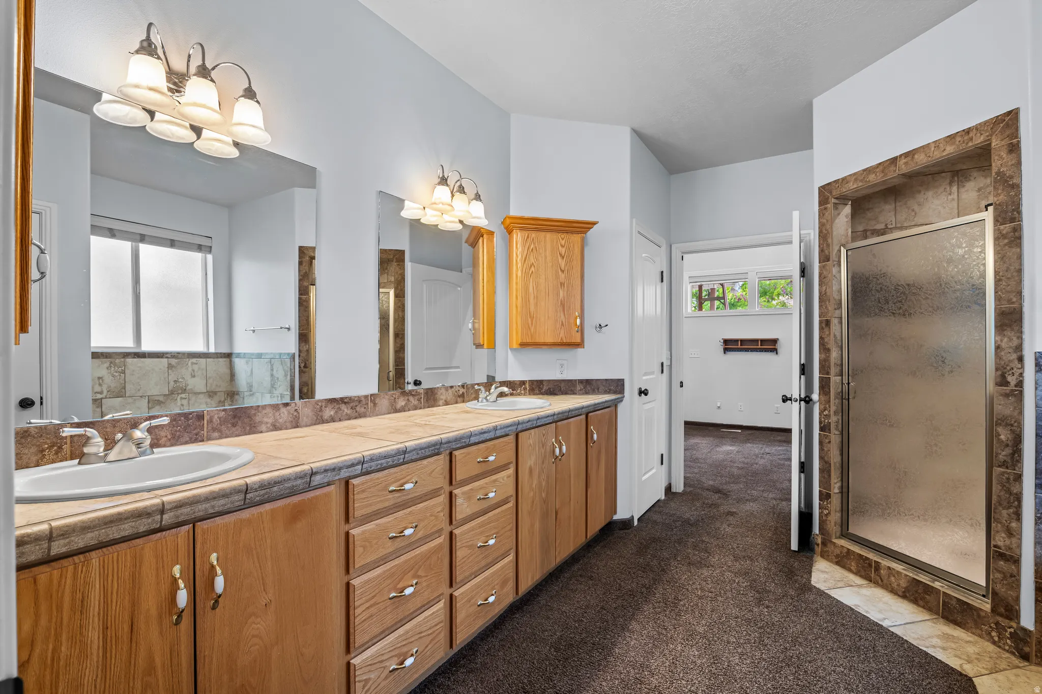 Spacious bathroom featuring a dual vanity with light wood cabinetry, tile countertops, and two integrated sinks