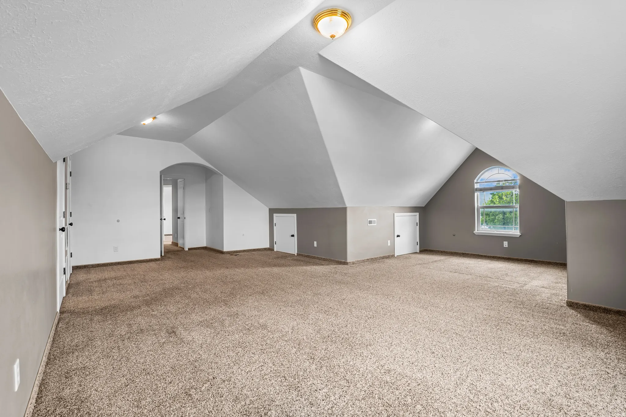 Spacious room featuring vaulted ceilings, neutral-toned carpeting, a prominent arched window, and multiple interior doors
