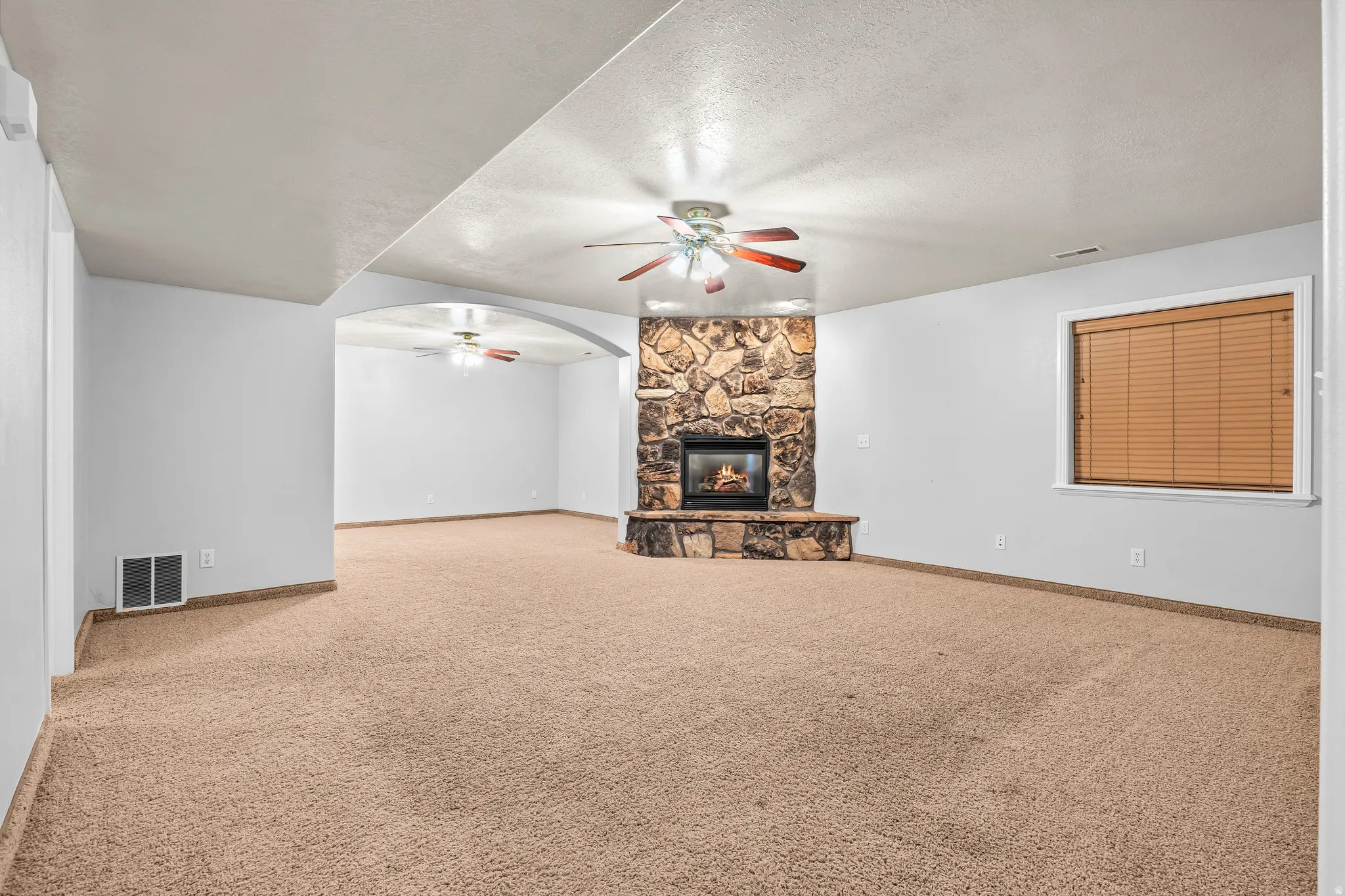 Expansive carpeted room featuring a stone fireplace with hearth, a ceiling fan, and a single window with blinds