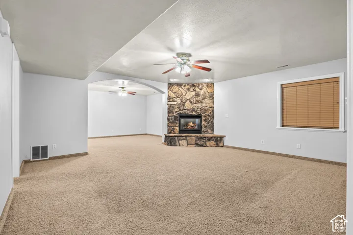 Expansive carpeted room featuring a stone fireplace with hearth, a ceiling fan, and a single window with blinds