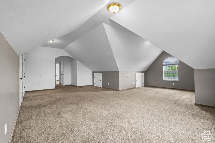 Spacious room featuring vaulted ceilings, neutral-toned carpeting, a prominent arched window, and multiple interior doors