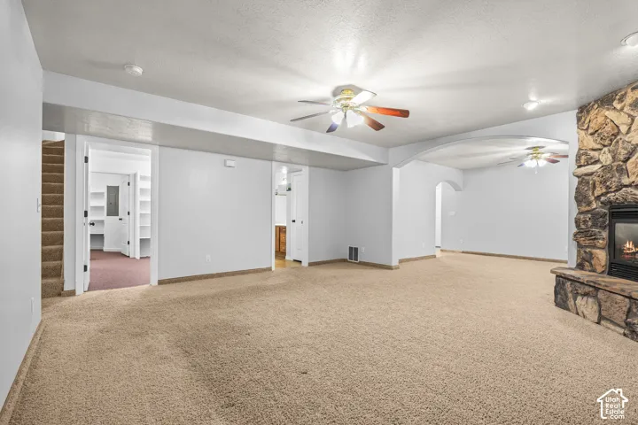 Spacious carpeted room featuring a stone-clad fireplace with a hearth, multiple ceiling fans, and an arched doorway leading to an additional space