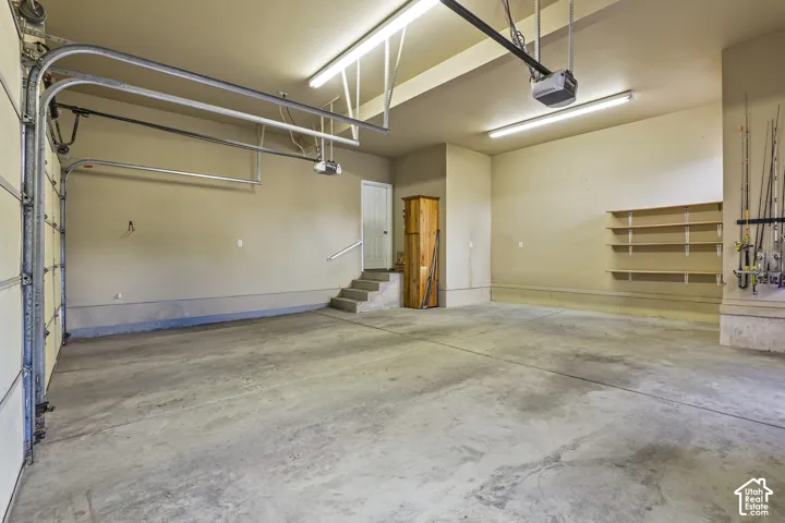 Spacious two-car garage featuring a concrete floor, built-in shelving, overhead fluorescent lighting, and a white interior access door with a handrail