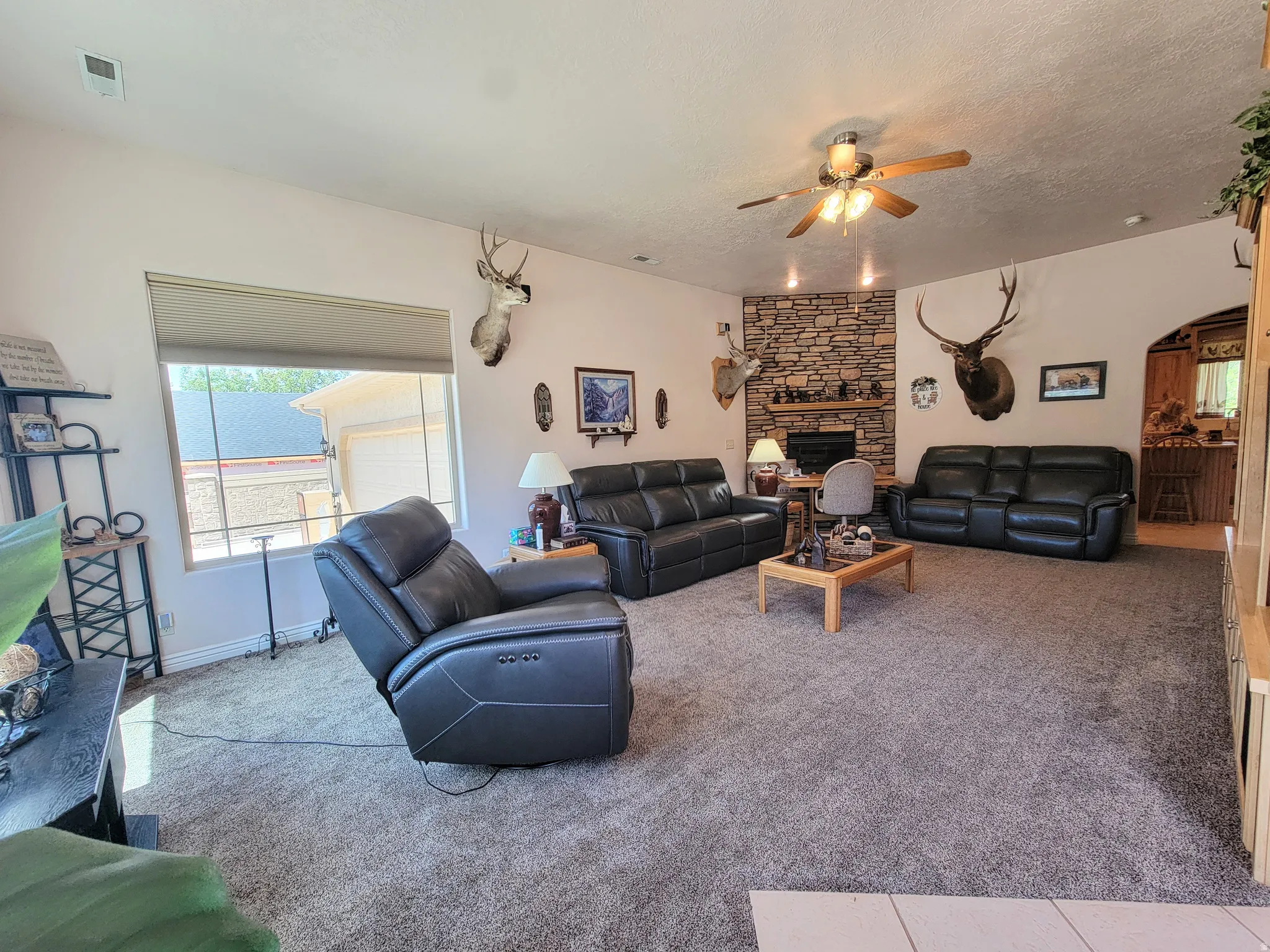 Spacious living area featuring a stacked stone fireplace with a wood mantle, a ceiling fan with integrated lighting, and carpet flooring