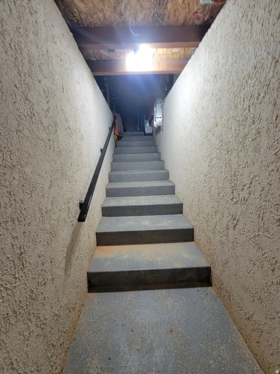 Concrete staircase with a black metal handrail, textured wall finish, and exposed ceiling joists
