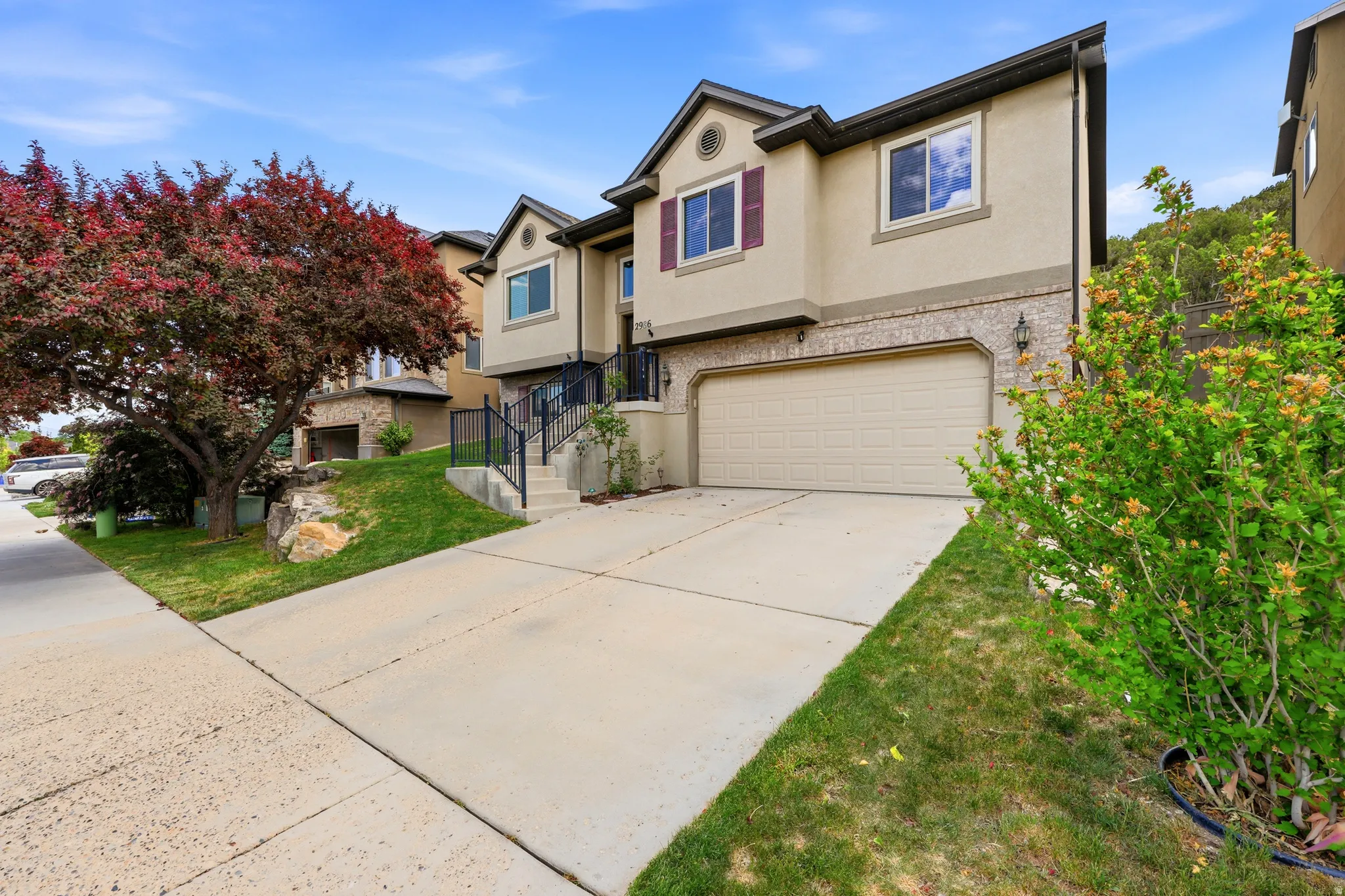 Stucco and brick exterior featuring a two-car garage, mature landscaping, concrete driveway, and front entry stairs with black railings