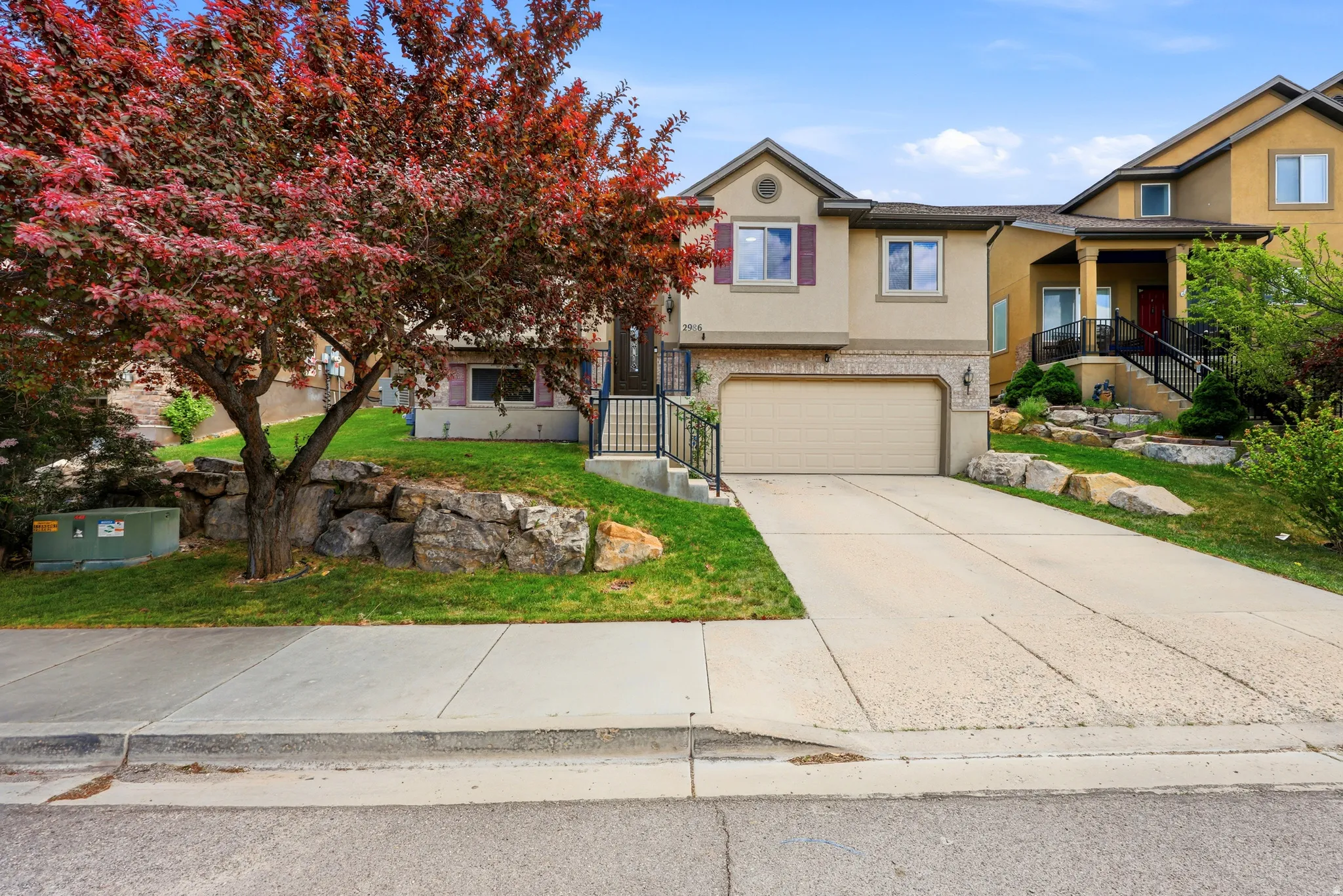 Two-story residence featuring a two-car garage, front yard landscaping with mature trees, a concrete driveway, and rockery accents