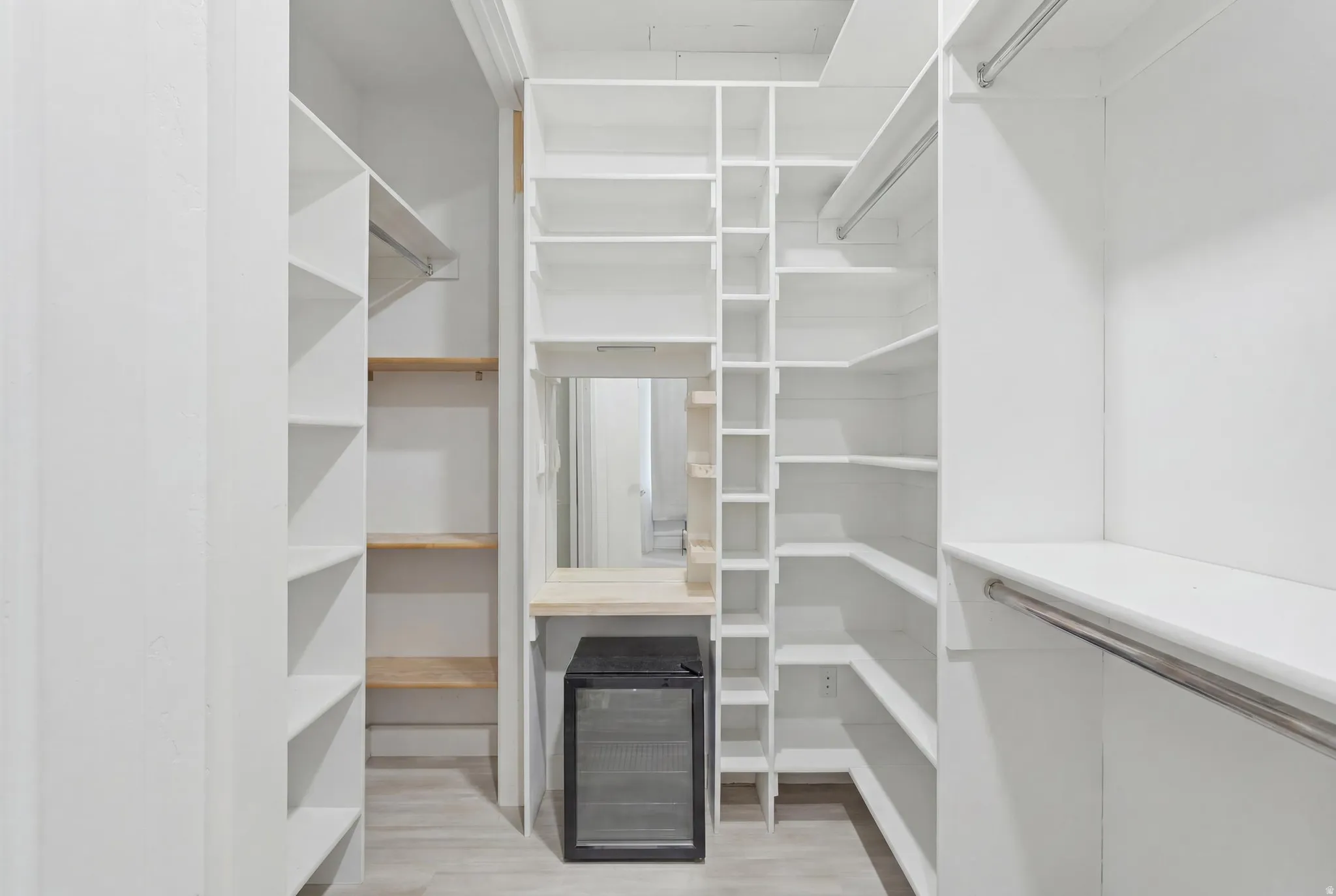 Custom walk-in closet featuring extensive white shelving, multiple hanging rods, corner shelving units, a built-in vanity with a mirror, and light wood-finish flooring
