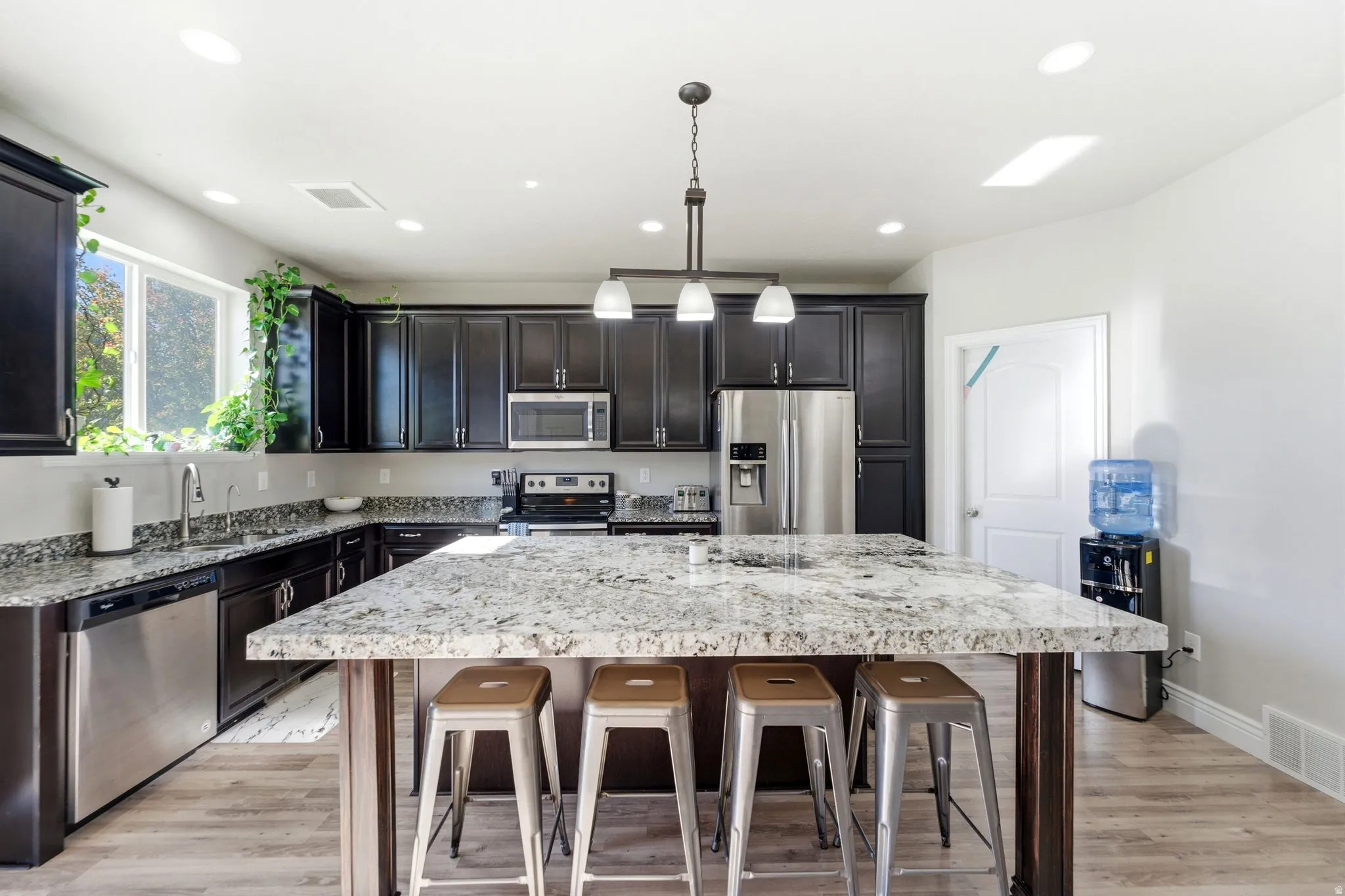 Spacious kitchen featuring an oversized island with a stone-finish countertop, dark wood-finish cabinetry, stainless steel appliances, recessed lighting, and wood-finish flooring