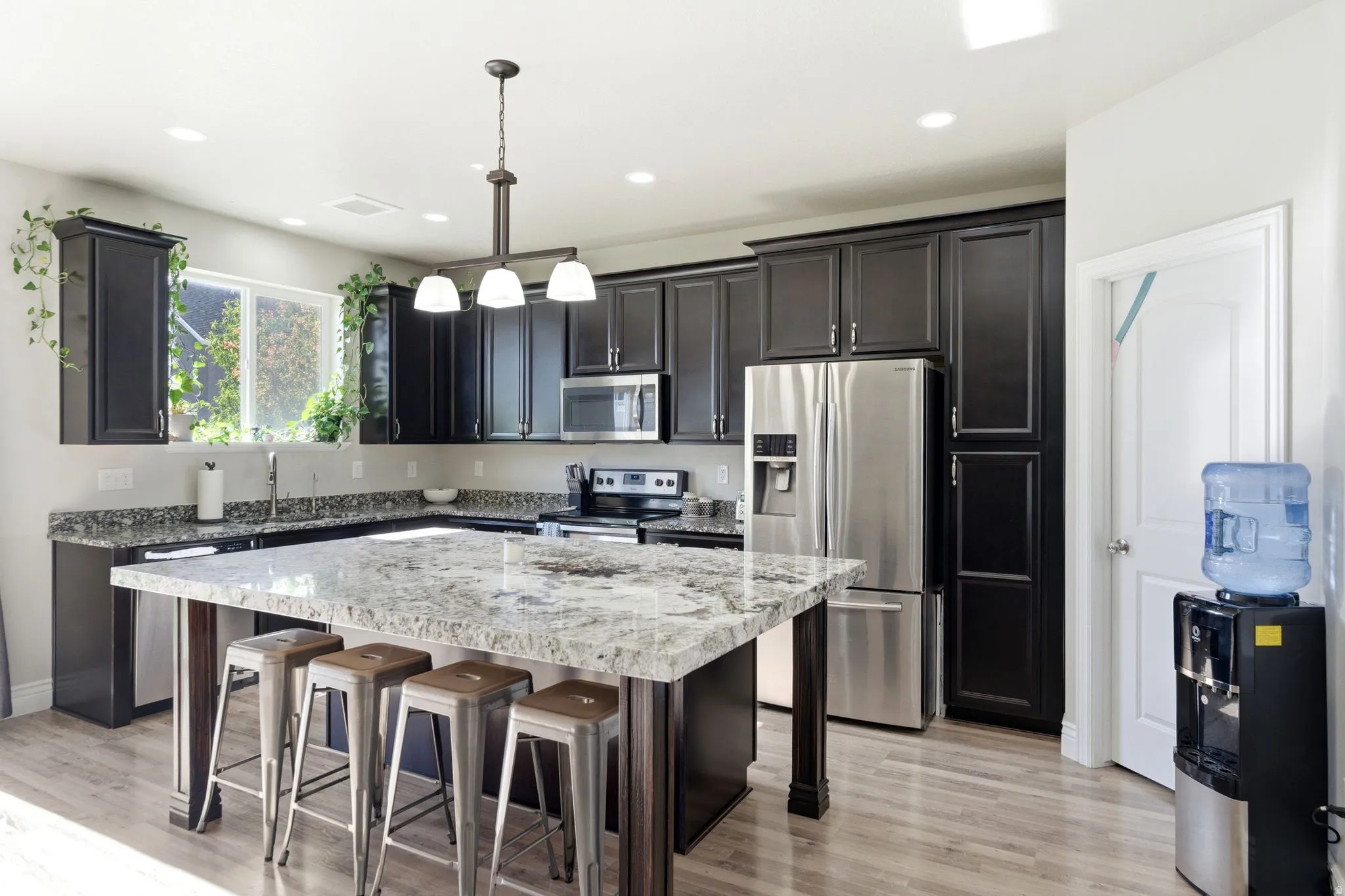 Spacious kitchen featuring an expansive island with a light-toned granite countertop, dark wood cabinetry, stainless steel appliances, wood-finish flooring, and recessed lighting