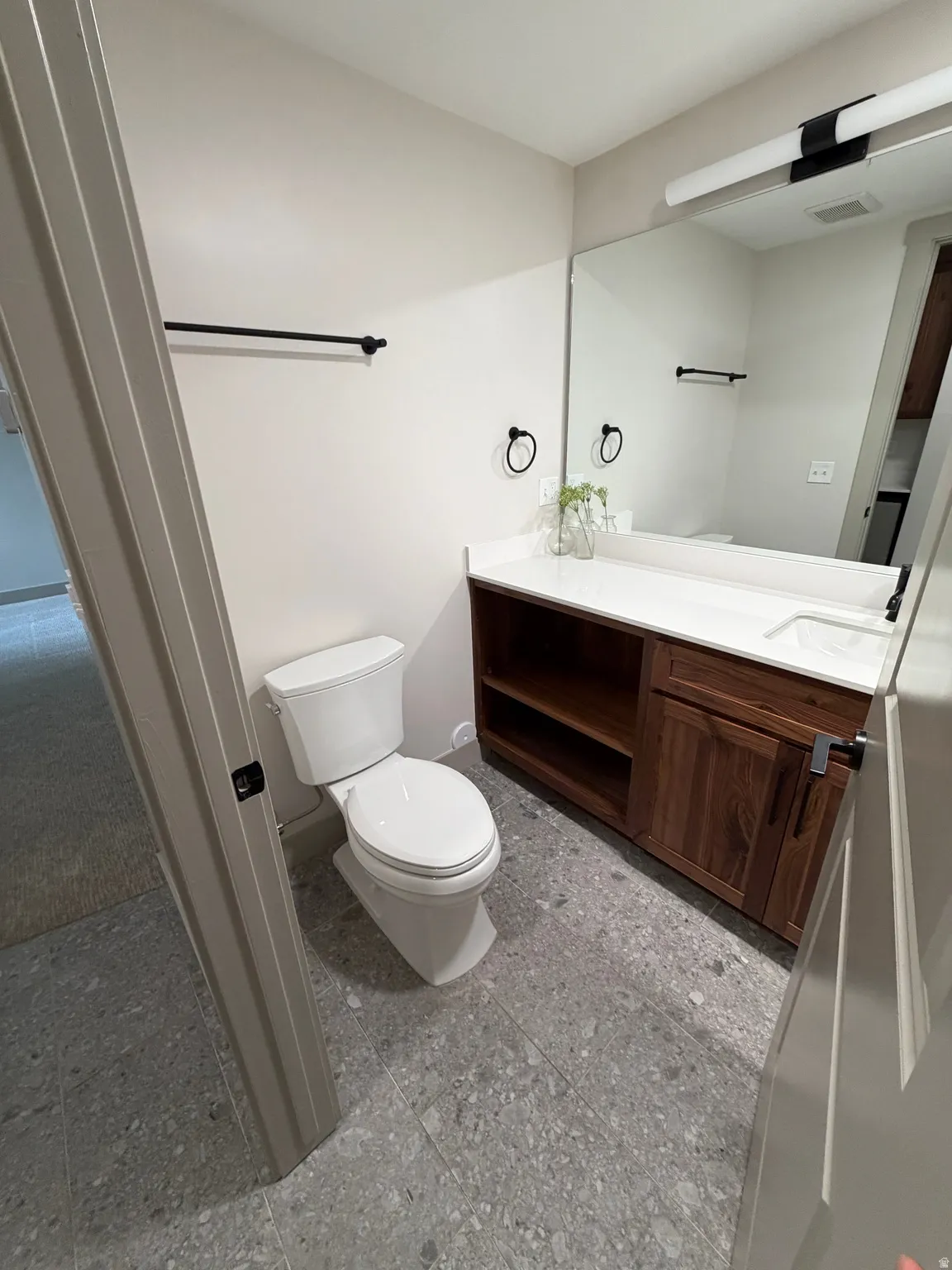 Bathroom featuring a white toilet, wood-finish vanity with open shelving and cabinet storage, white countertop with integrated sink, and large wall-mounted mirror with linear lighting fixture