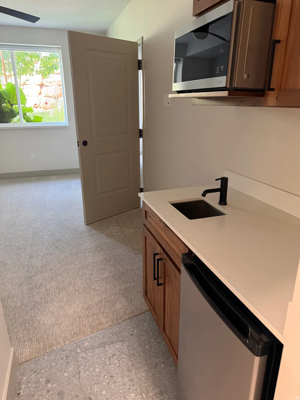 Wet bar featuring a stainless steel microwave, wood-finish cabinetry with black hardware, white countertop with an undermount sink and matte black faucet, and a stainless steel compact refrigerator