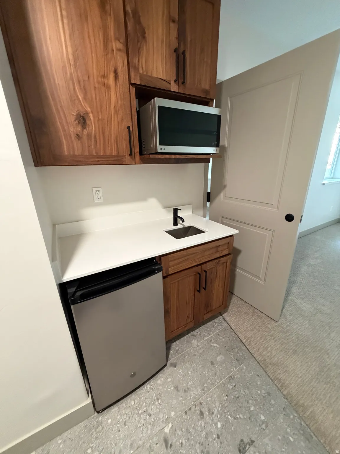 Built-in wet bar featuring rich wood-finish cabinetry, a stainless steel microwave, a compact refrigerator, a contemporary sink, and a sleek black faucet, all set against a white countertop